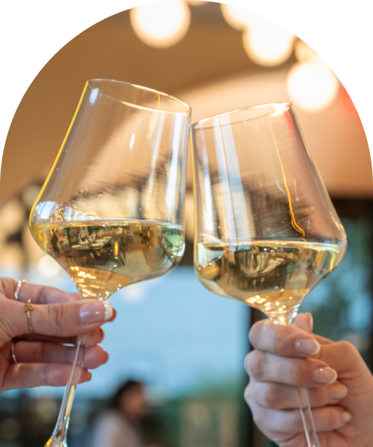 Two hands toast with glasses of white wine against a blurred, warm-lit background.