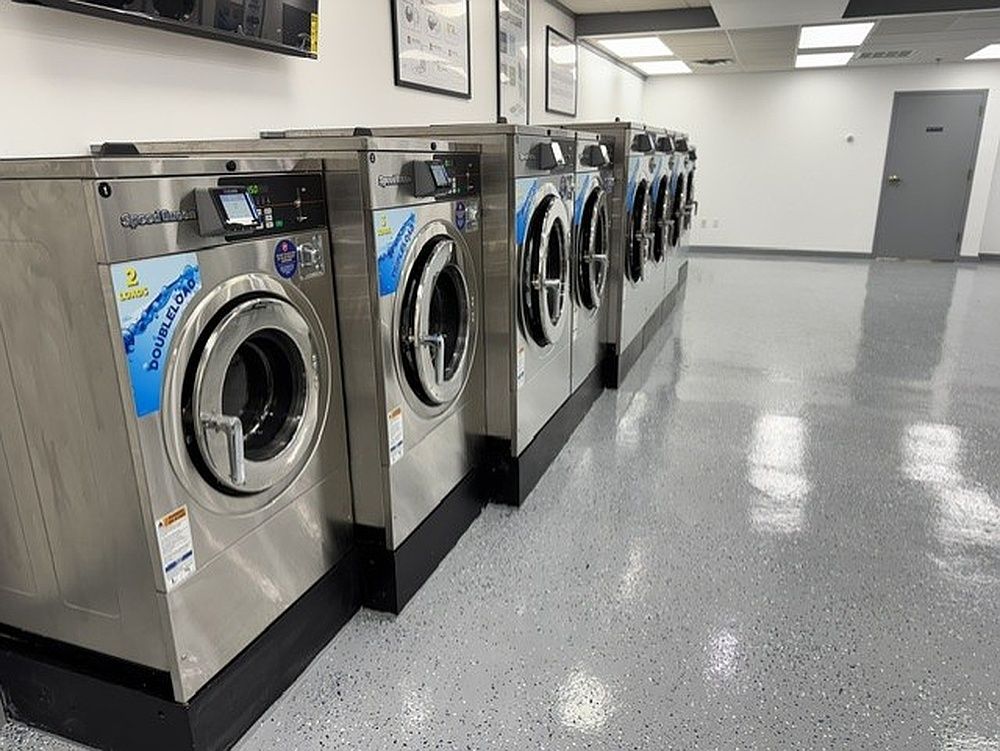 Row of silver front-loading washing machines in a clean laundromat with gray speckled floors and white walls