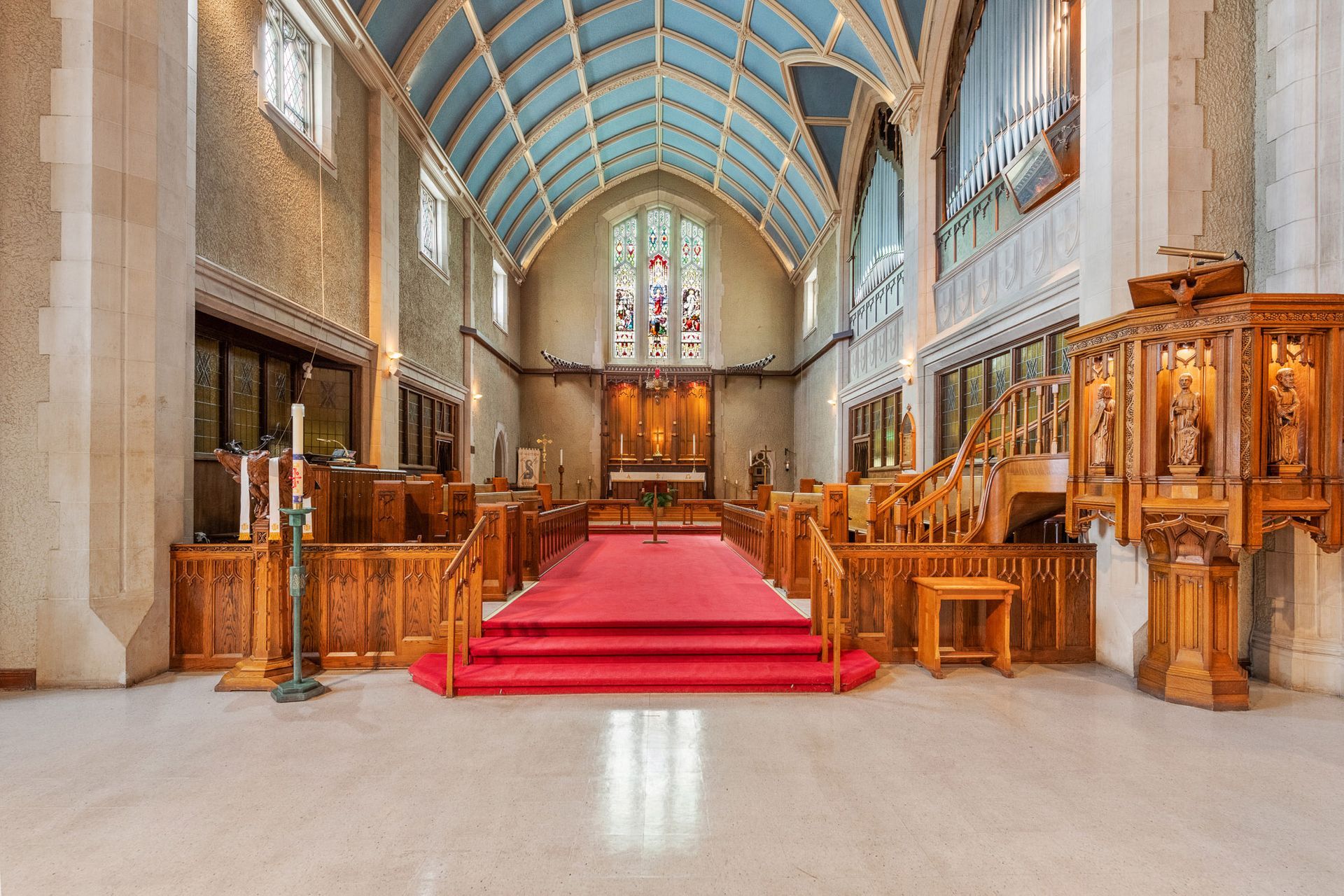 Church interior with red-carpeted altar, wooden pews, stone columns, and blue arched ceiling
