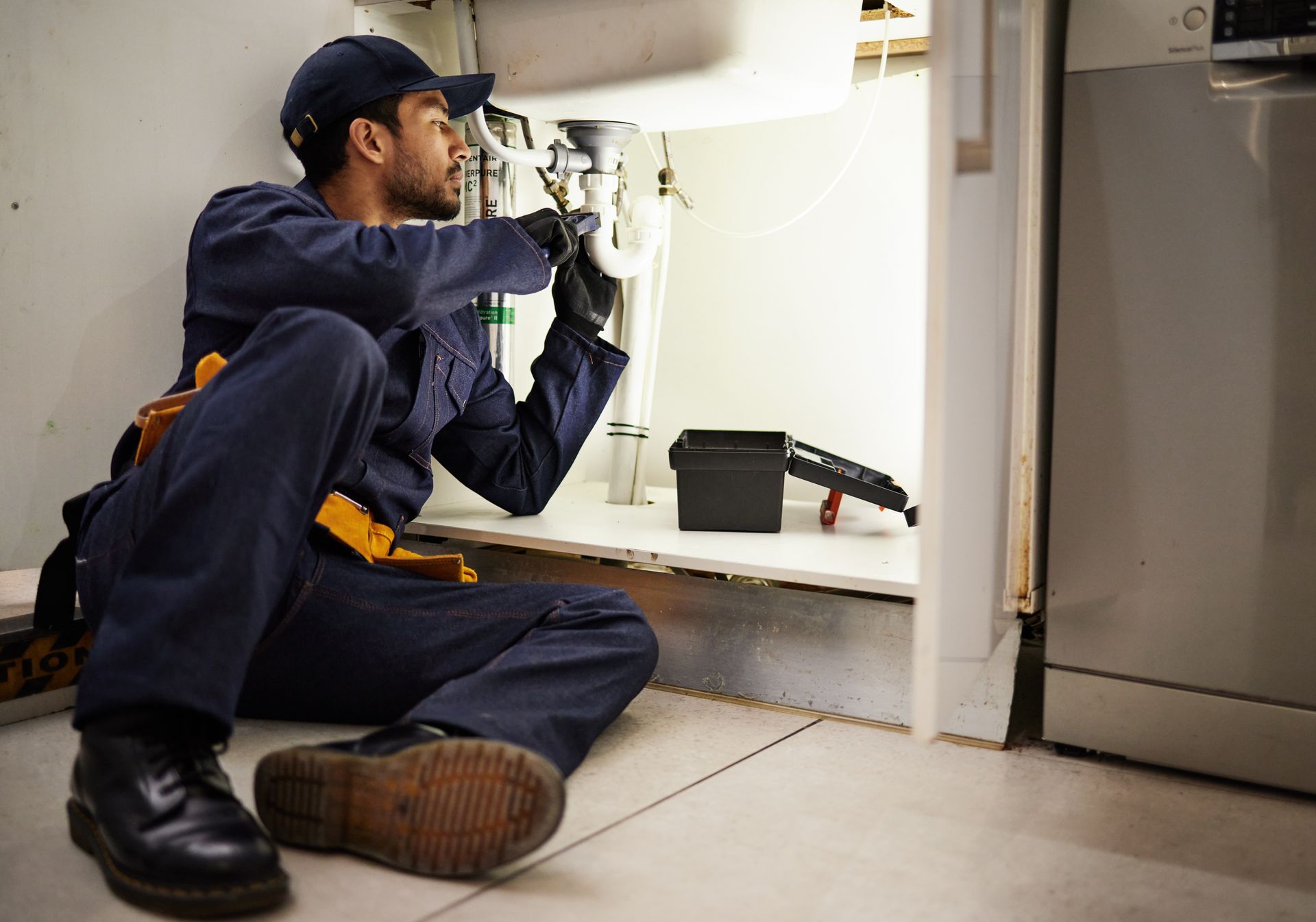 A plumber sitting on the floor fixing a sink with tools and a light after an emergency.