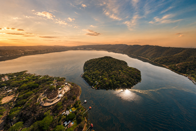 Vista aérea de una puesta de sol sobre el lago coatepeque tranquilo con una pequeña isla verde, rodeado de colinas y un cálido cielo anaranjado.
