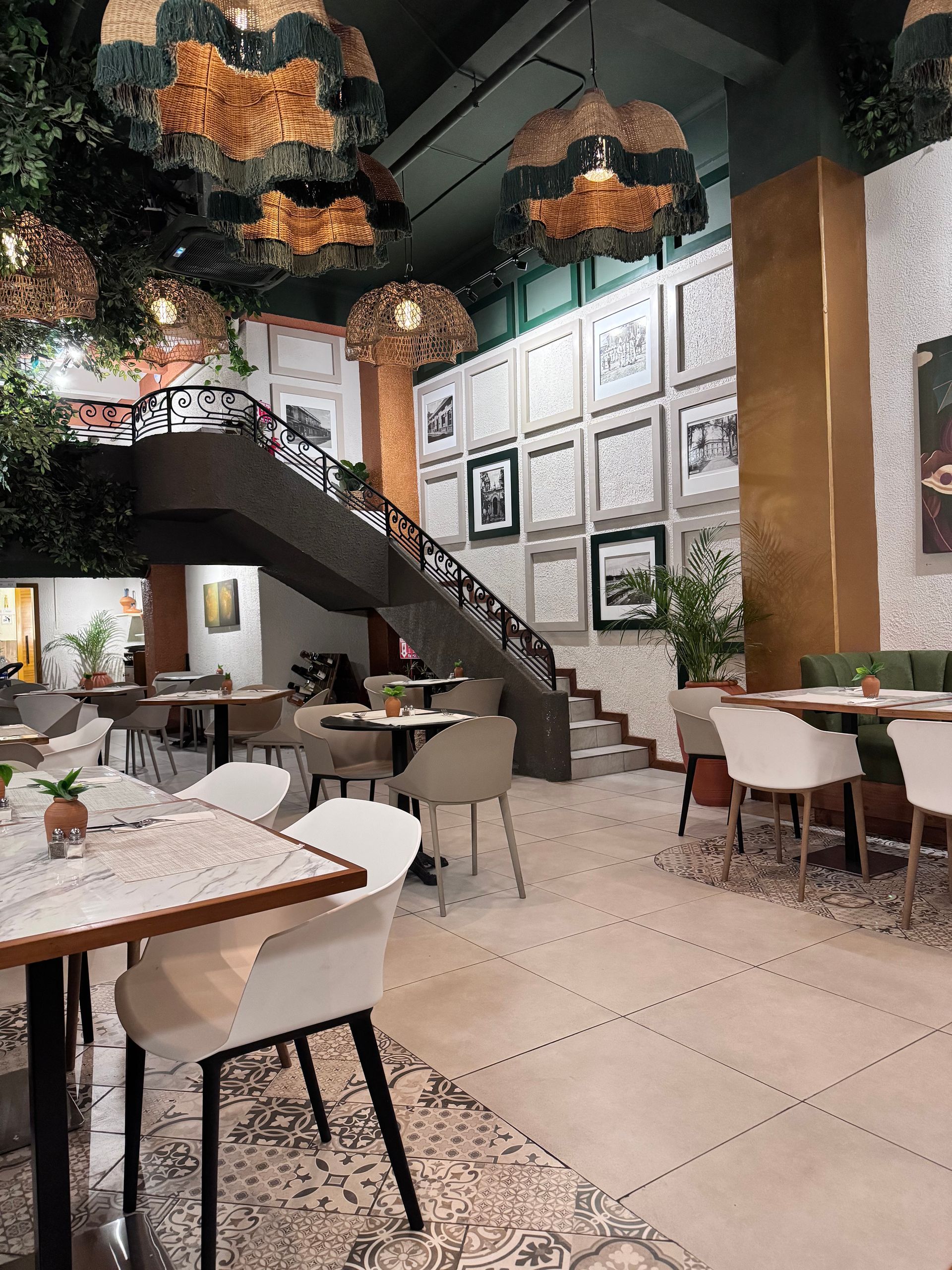 Interior de cafetería moderna con mesas, sillas, escalera y luces colgantes cálidas.