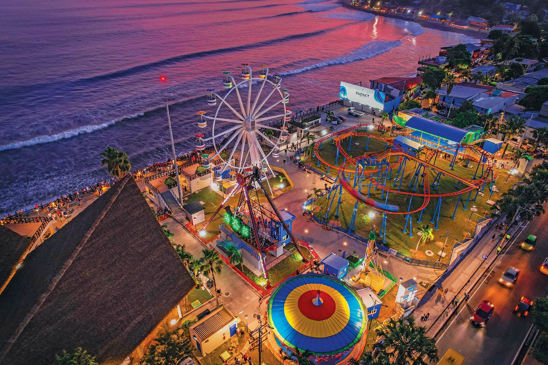 Vista aérea de un colorido parque de atracciones junto al mar con noria, juegos mecánicos y luces brillantes al anochecer.