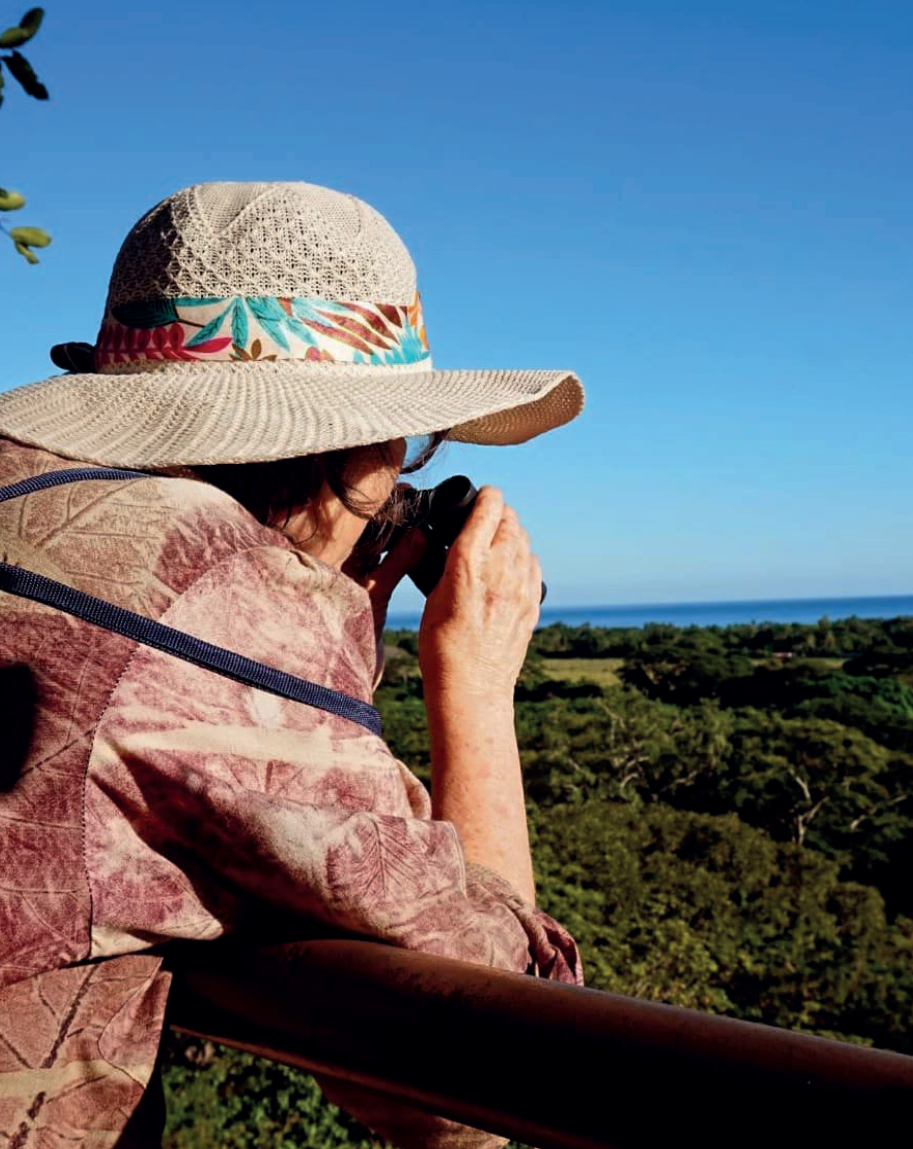 Una persona con un sombrero de paja fotografía un paisaje verde y el océano azul desde un balcón.