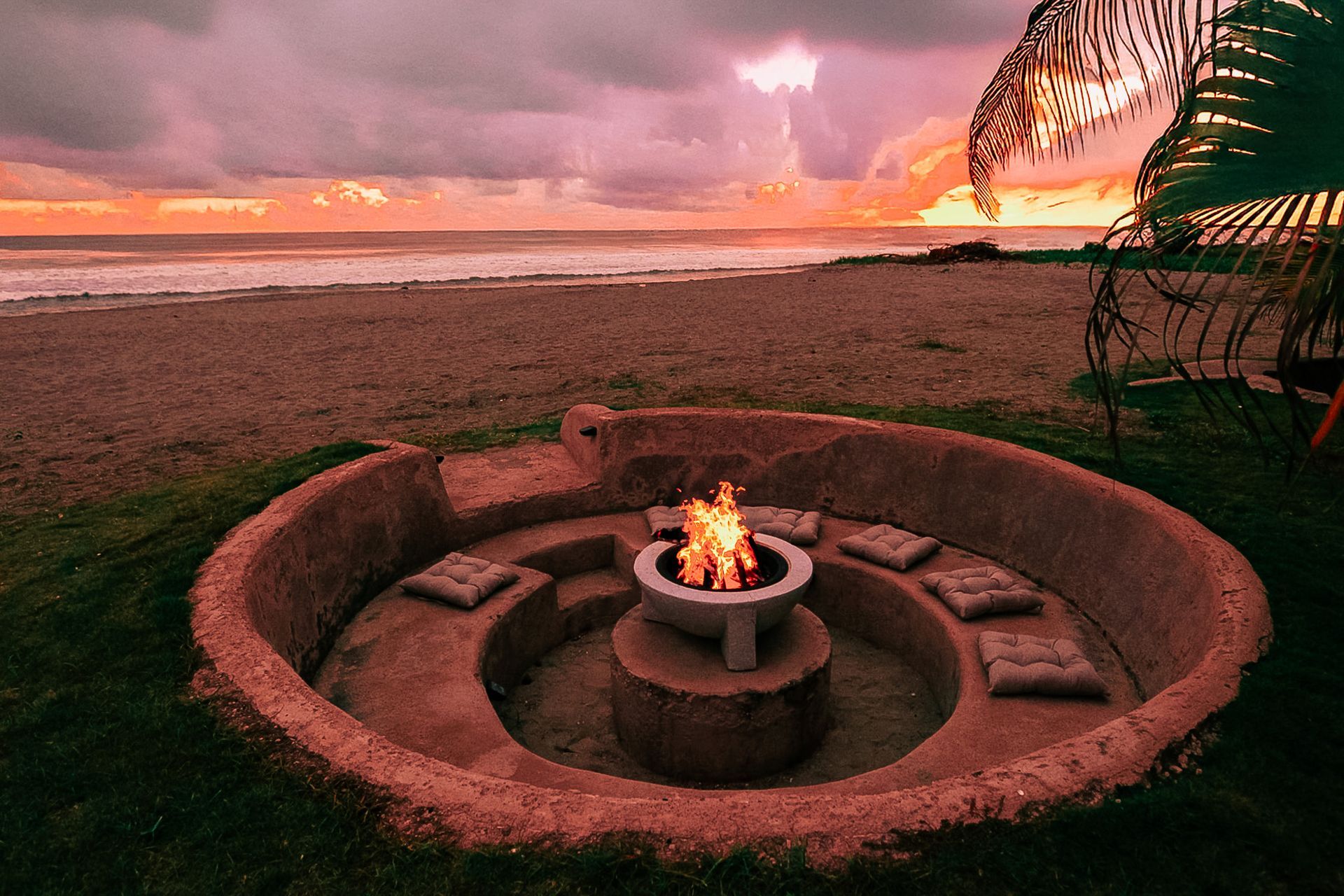 Hoguera circular de piedra en una playa al atardecer, con llamas brillantes bajo un cielo rosa anaranjado.