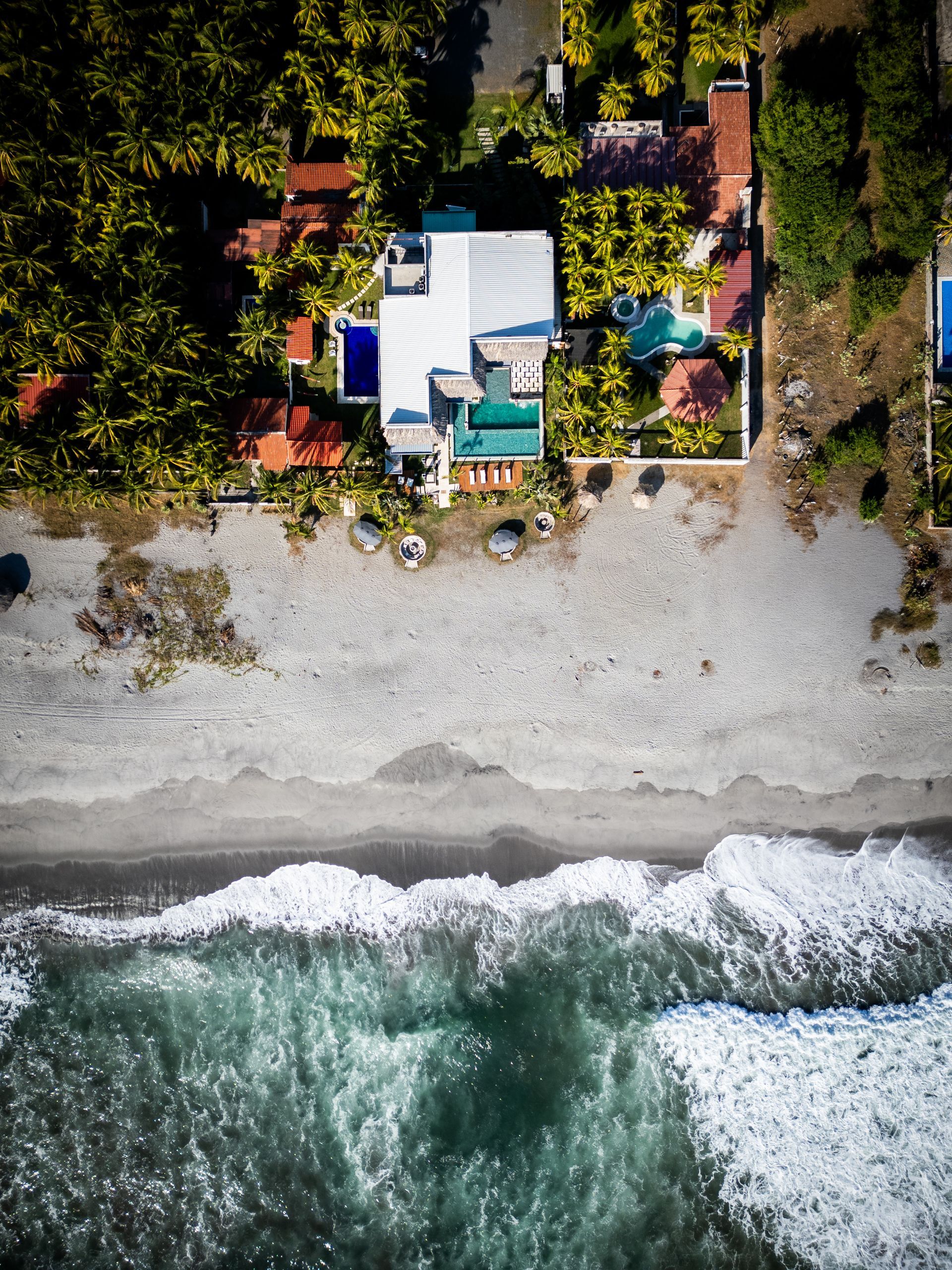 Vista aérea de cardedeu cangrejera el salvador frente al mar con un patio blanco, una orilla de guijarros y olas rompiendo abajo.