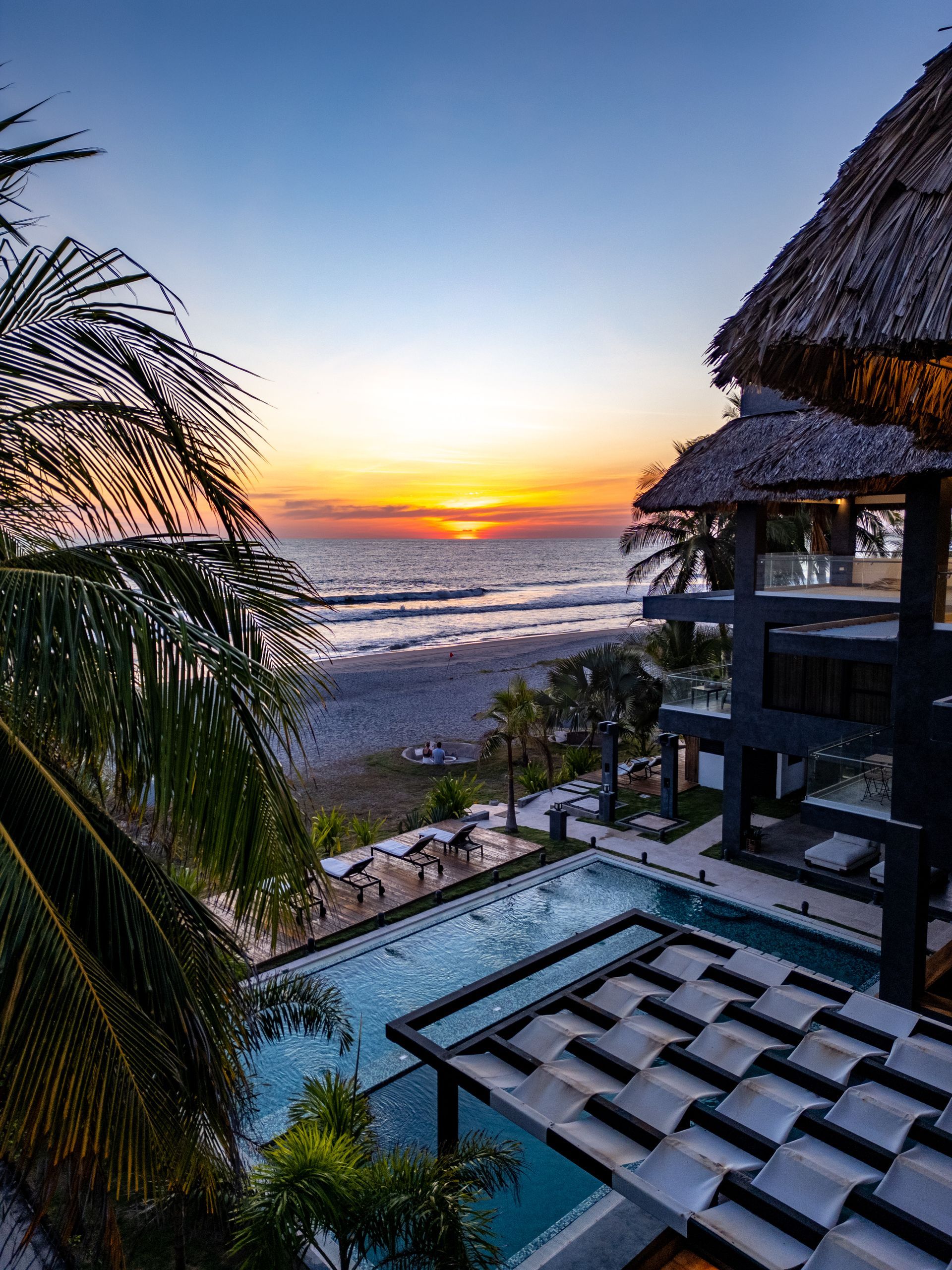 Piscina en cardedeu cangrejera el salvador frente al mar al atardecer, con palmeras, edificios con techos de paja y el mar al fondo.