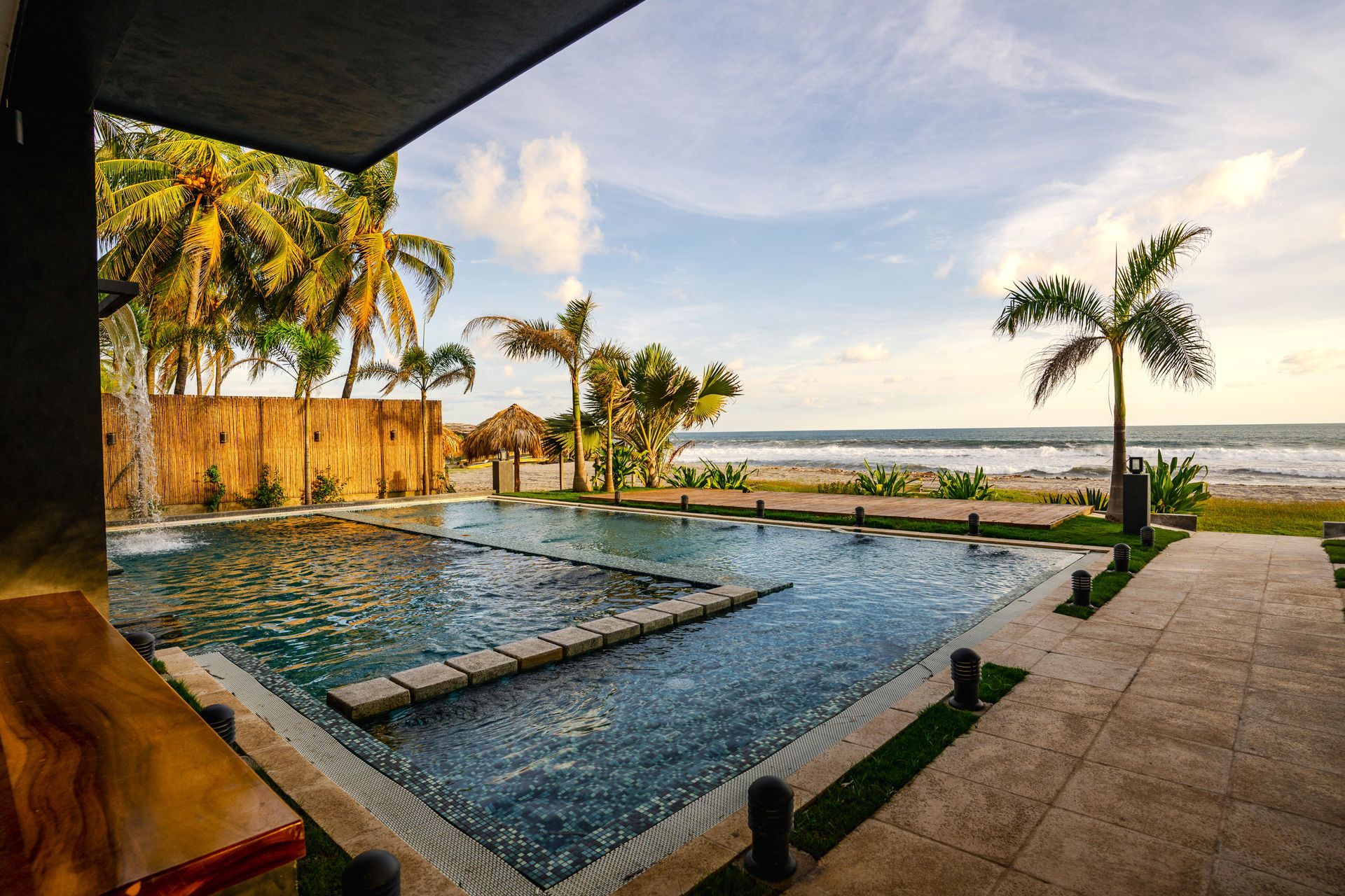 Piscina infinita junto a una playa tropical al atardecer, enmarcada por palmeras y un patio sombreado en cardedeu cangrejera el salvador