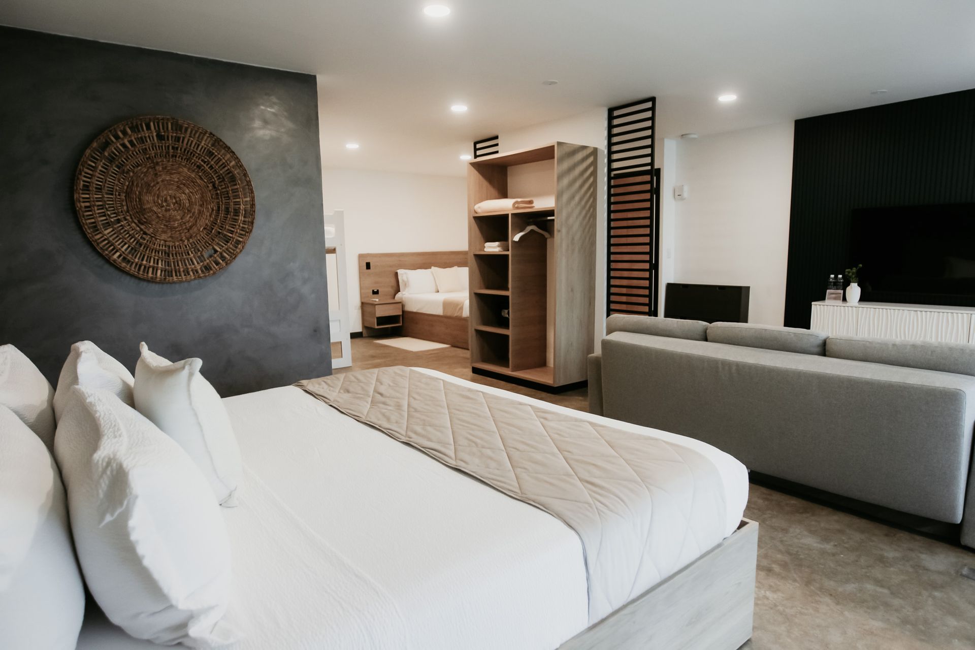 Dormitorio moderno con cama blanca, pared de acento oscura y estanterías abiertas junto a una sala de estar con sofá gris.