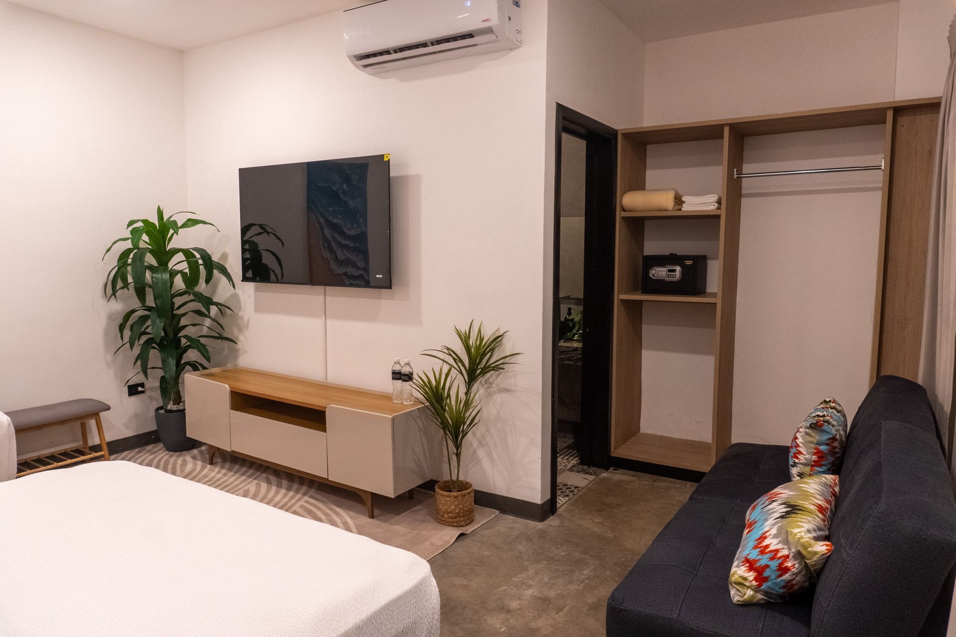 Dormitorio moderno con televisor de pared, cama, planta, armario abierto y sofá cerca de un aire acondicionado.