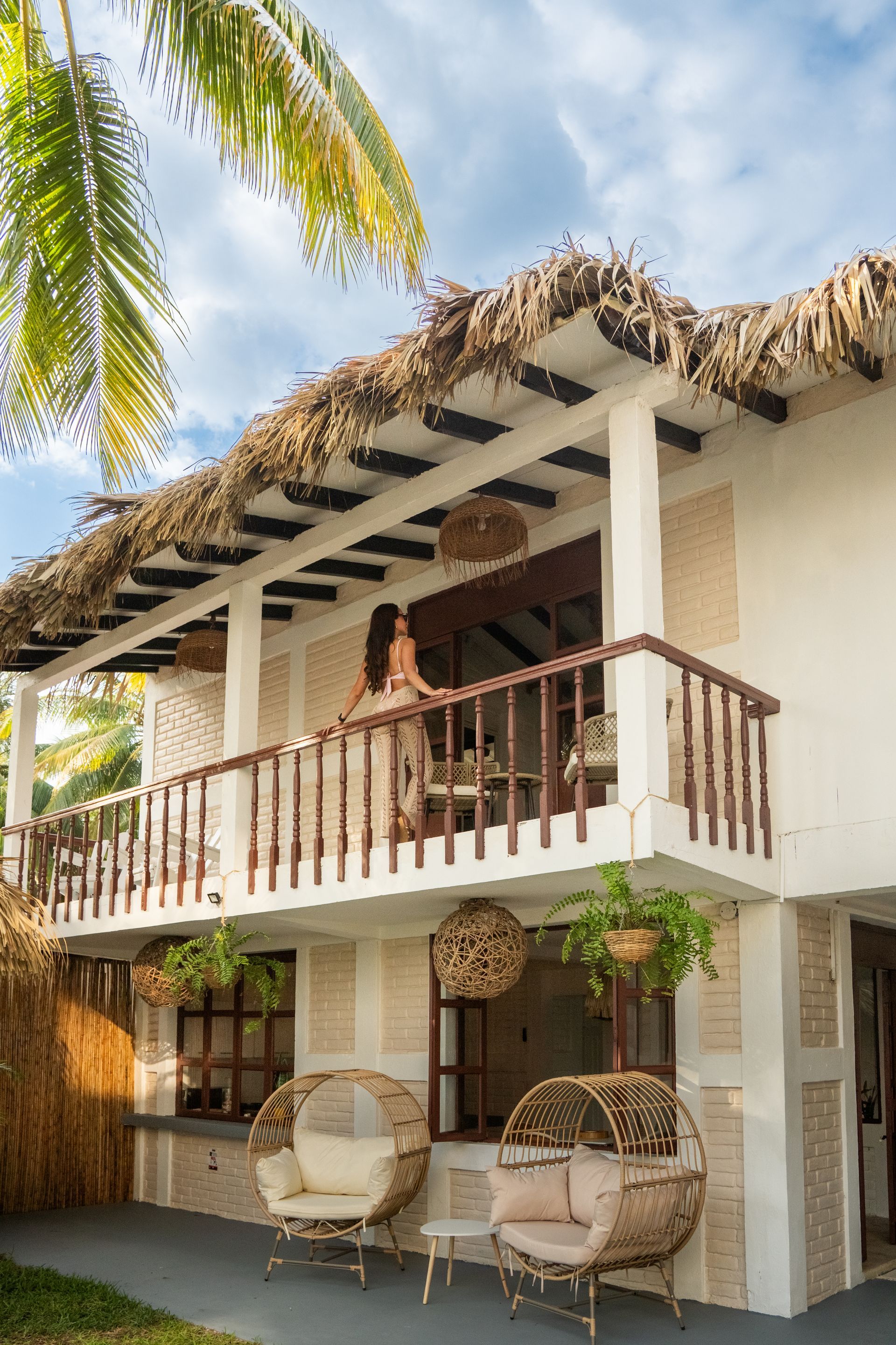 Villa tropical de dos plantas con techo de paja, balcón y sillas de mimbre bajo una palmera.