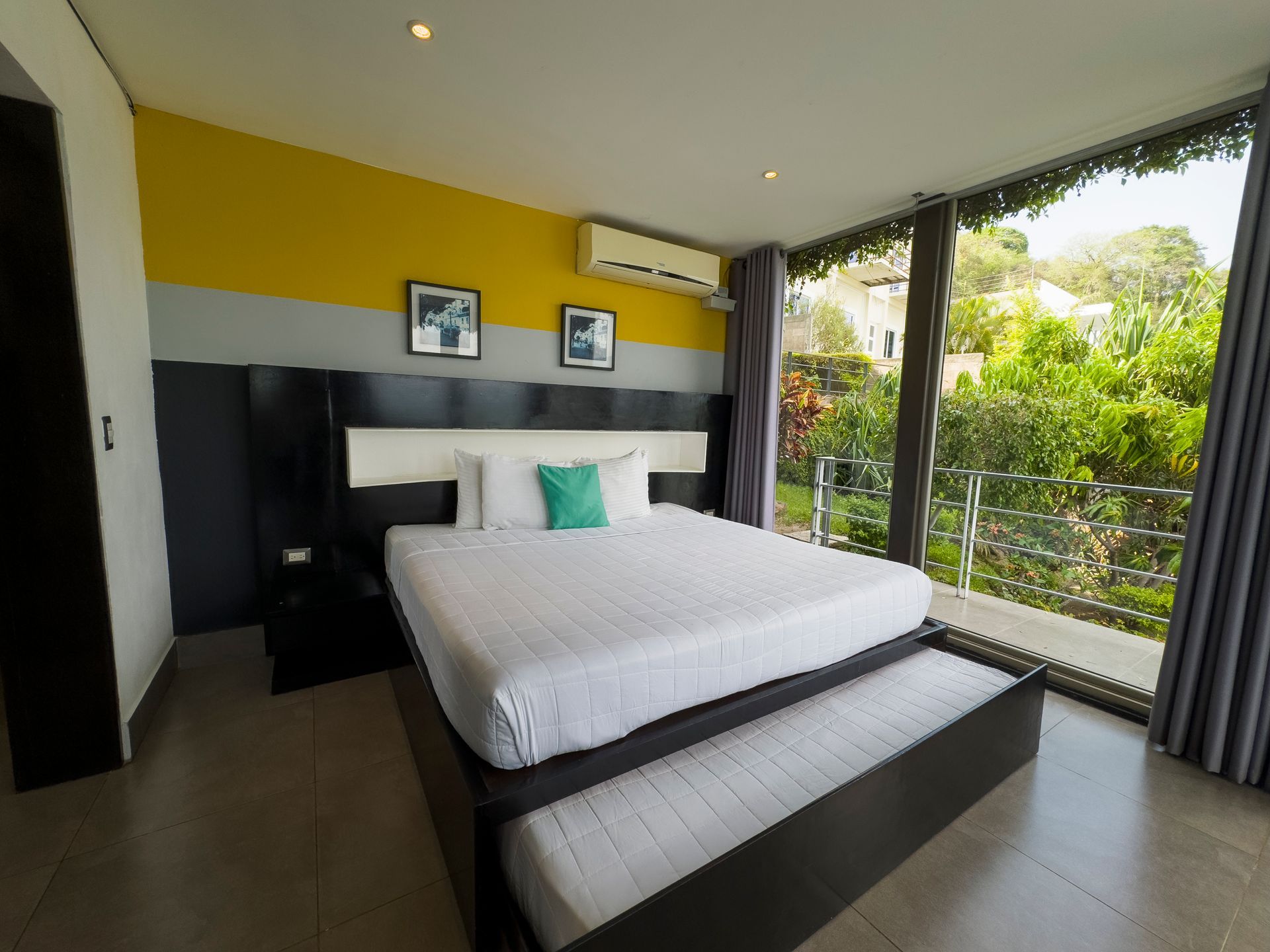 Dormitorio moderno con una cama grande, una pared decorativa amarilla y una ventana que va del suelo al techo con vistas a la vegetación.