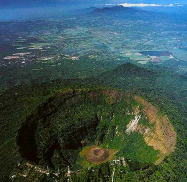 Vista aérea de un cráter volcánico con laderas verdes y un cono central, con vistas a campos y montañas a lo lejos.