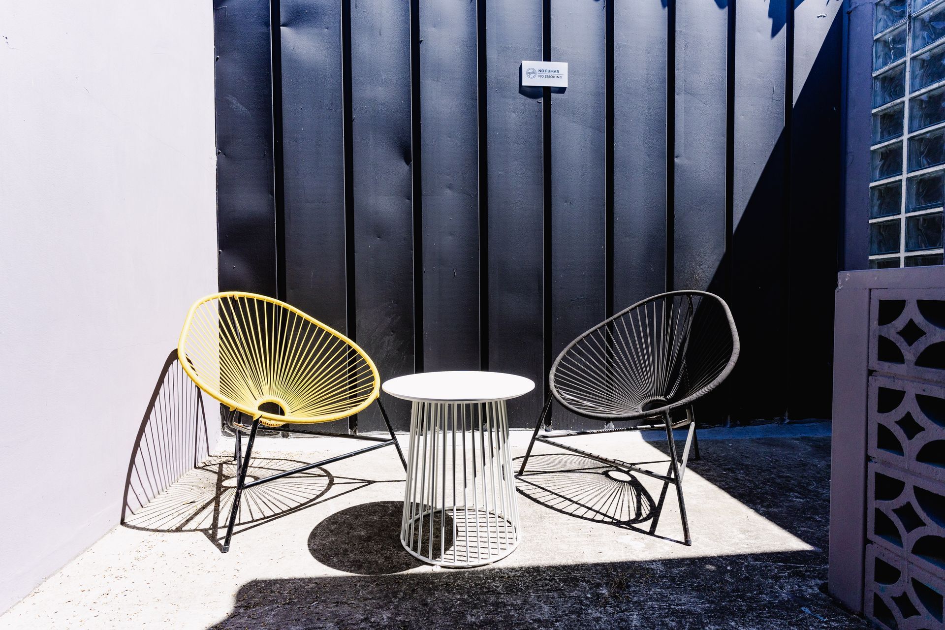 Dos sillas modernas de alambre y una pequeña mesa redonda en un patio soleado, frente a una pared vertical oscura en hotel cardedeu express