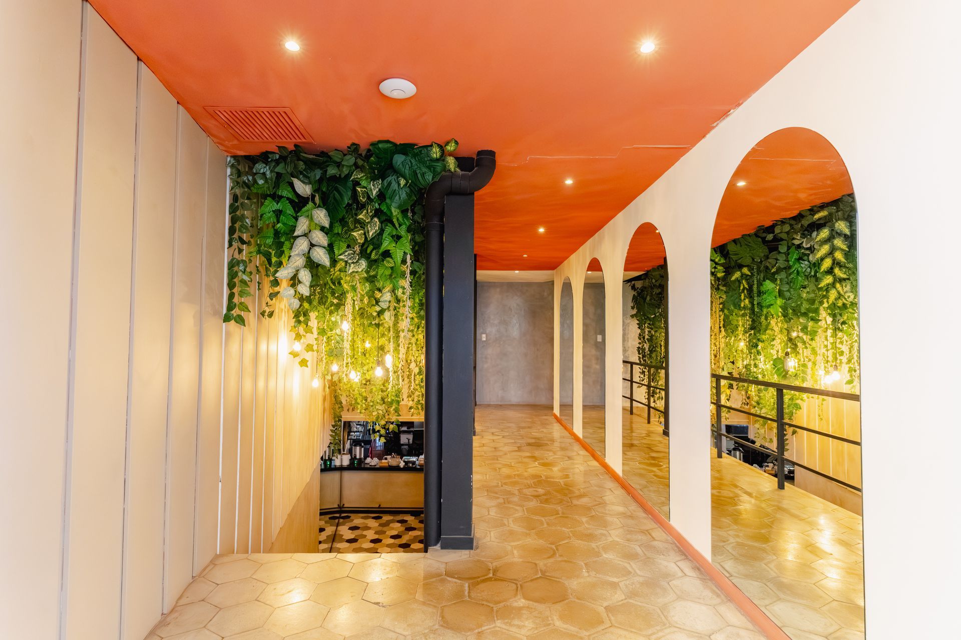 Pasillo interior moderno con techo naranja, arcos blancos y plantas verdes colgantes.