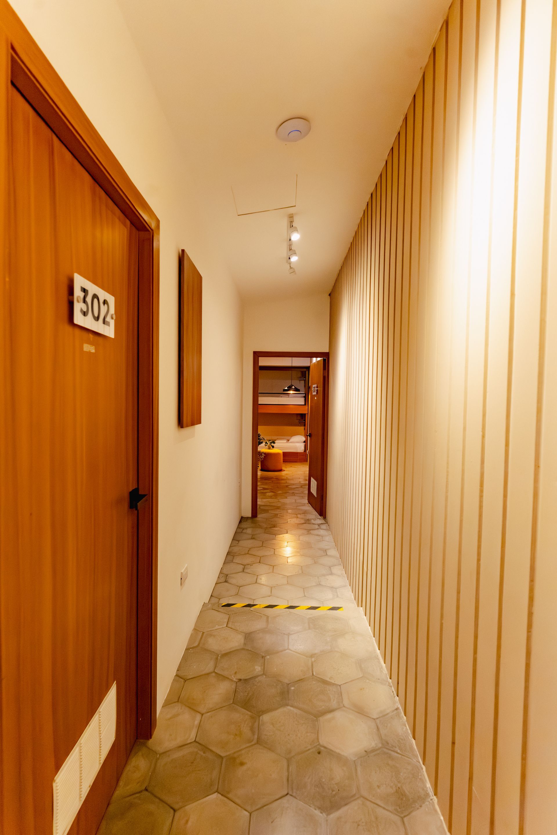 Pasillo estrecho con puertas de madera, suelo de baldosas hexagonales e iluminación cálida que conduce a una puerta.