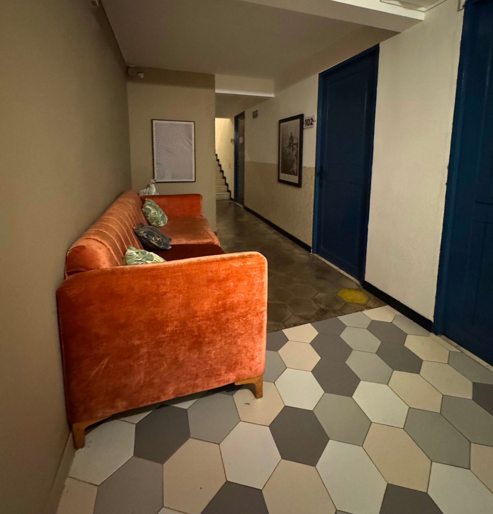 Salón-pasillo con sofá naranja, suelo de baldosas estampadas, puertas azules y cuadros enmarcados.