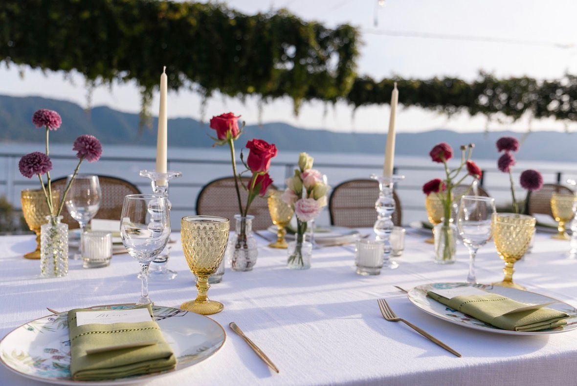 Elegante mesa de comedor al aire libre con mantel blanco, copas doradas, velas y centros de mesa florales rosas junto al agua en el hotel cardedeu coatepeque