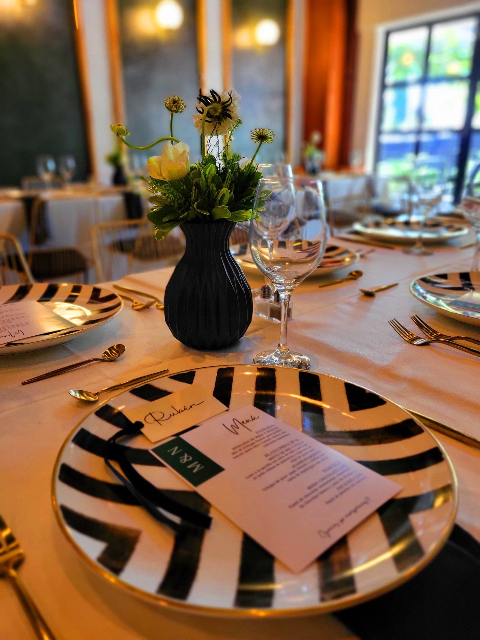 Elegante mesa de comedor puesta con platos a rayas, menús plegados, copas de vino y un pequeño centro de mesa floral en silvestre centro historico el salvador