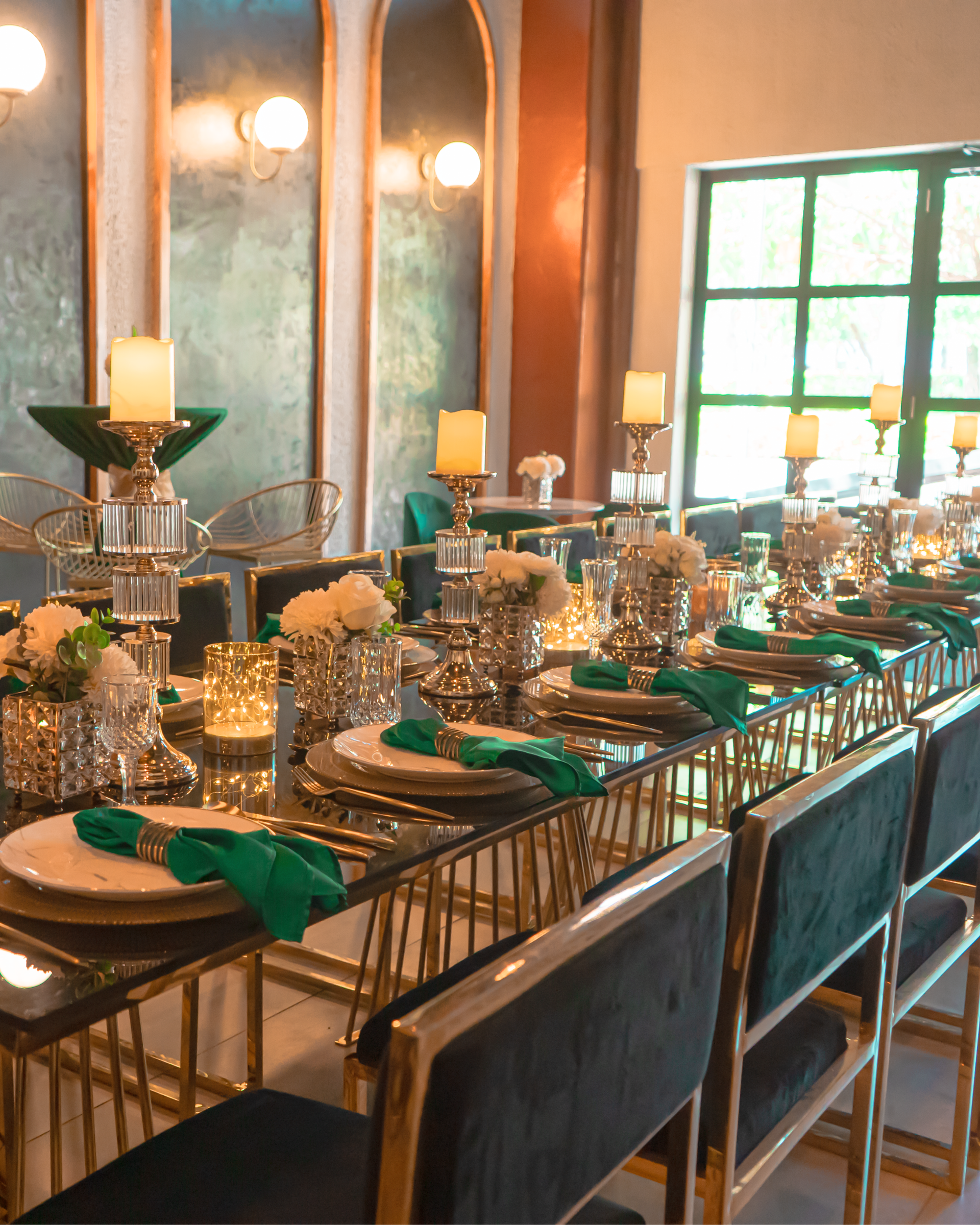 Elegante mesa de comedor con detalles dorados, servilletas verdes, velas y cristalería en un luminoso restaurante.