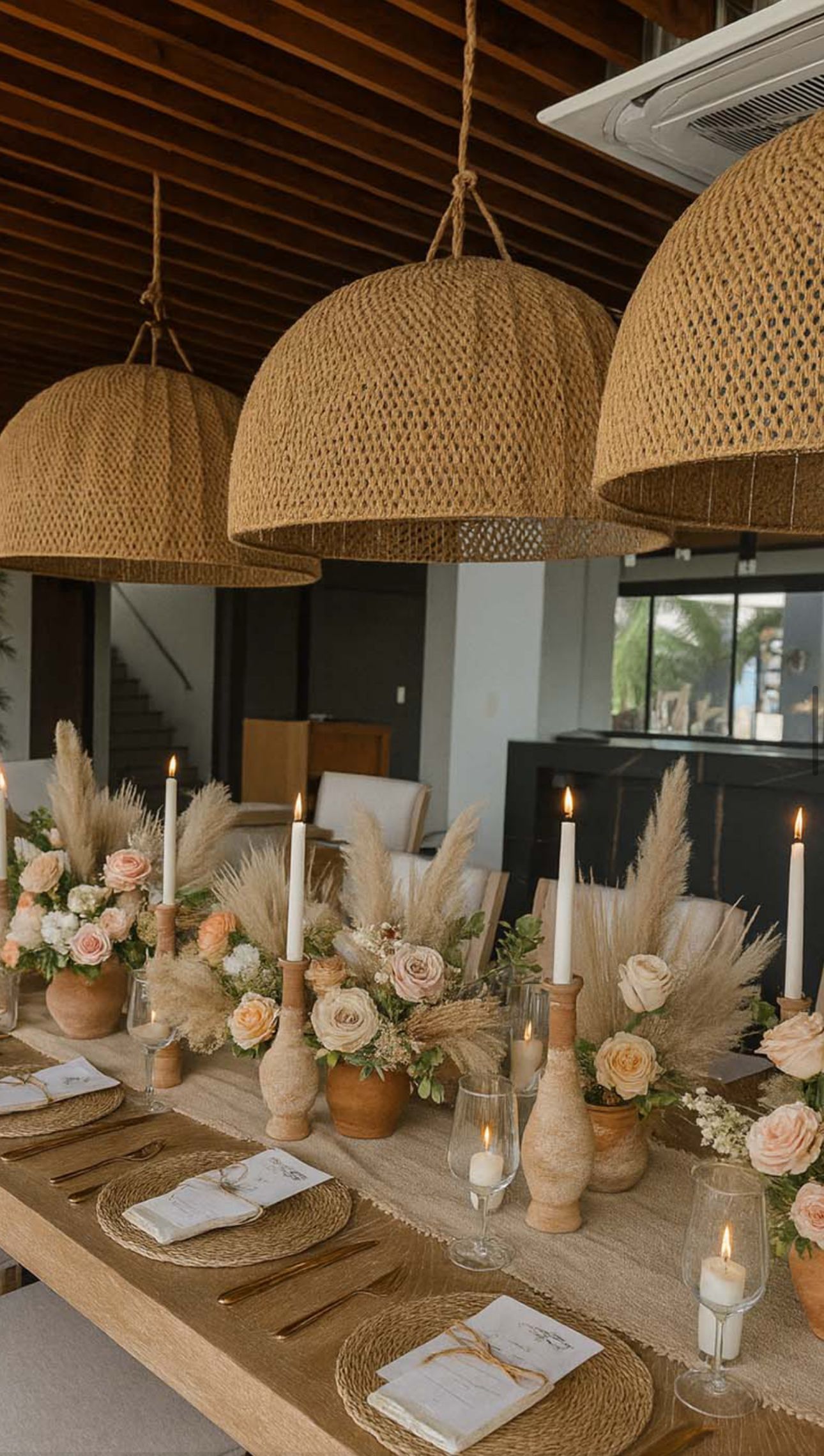 Mesa de comedor decorada bajo lámparas colgantes tejidas, con arreglos florales, velas y vajilla en cangrejera cardedeu
