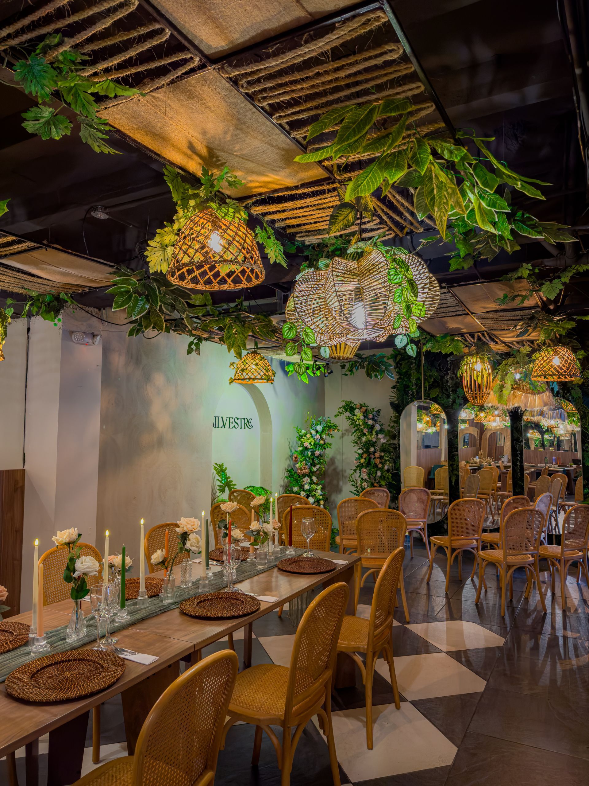 Interior del restaurante con iluminación cálida, sillas de mimbre, mesas de madera, plantas y lámparas colgantes en silvestre san benito