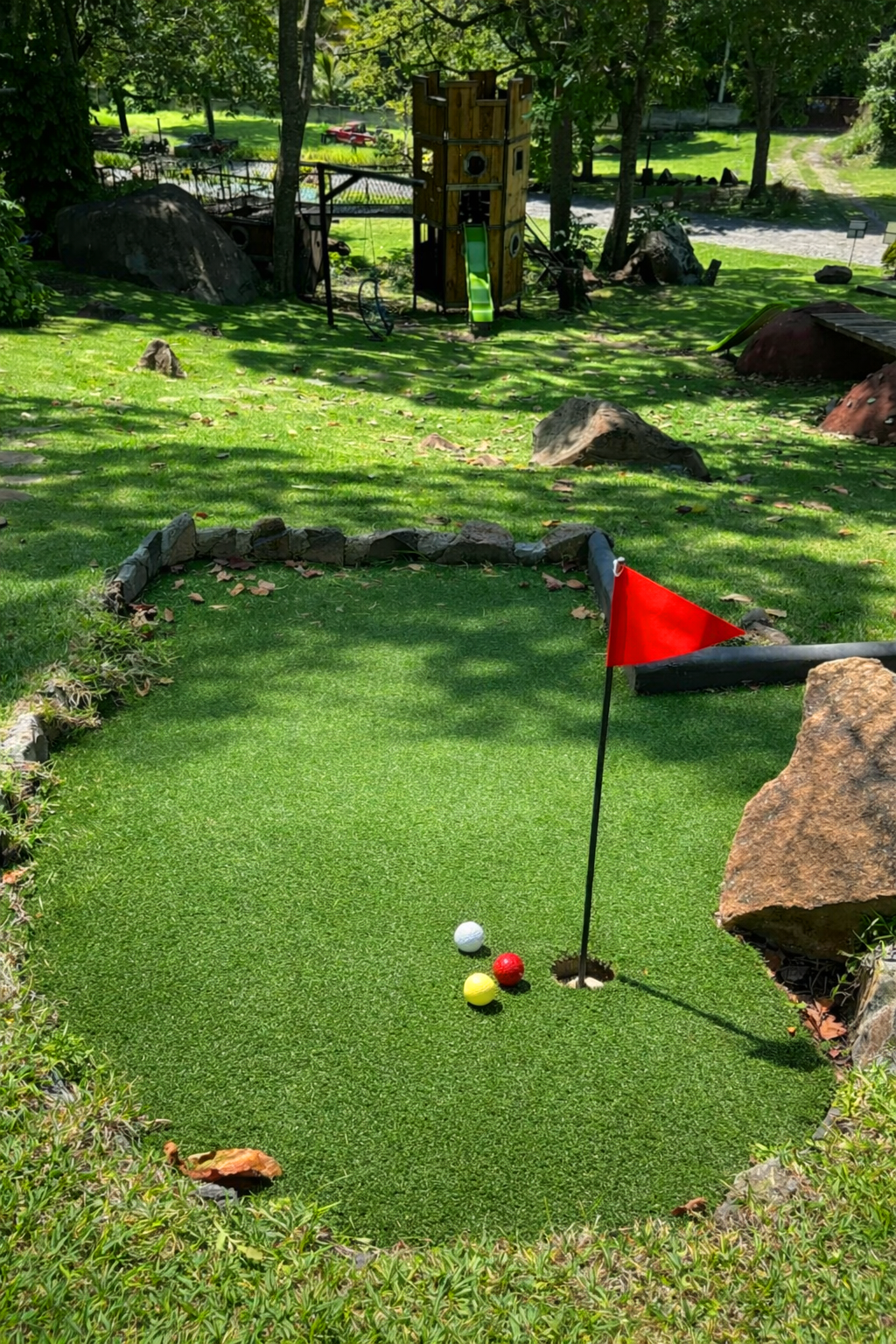 Campo de minigolf con bandera roja y tres pelotas de golf cerca de una roca, rodeado de árboles y rocas.