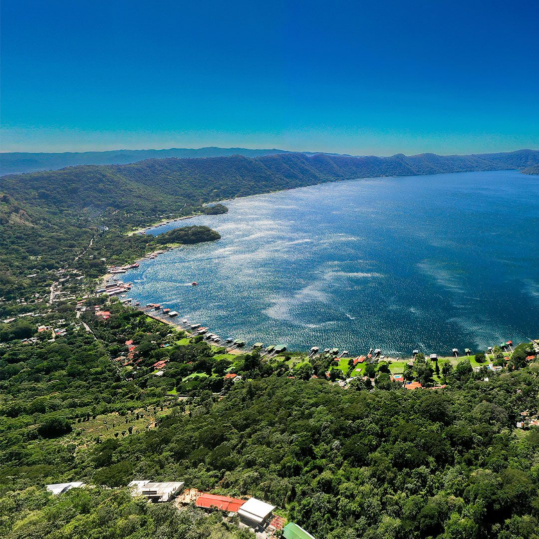 Vista aérea de una bahía azul bordeada de exuberantes colinas verdes y un pueblo costero bajo un cielo despejado en el lago coatepeque el salvador