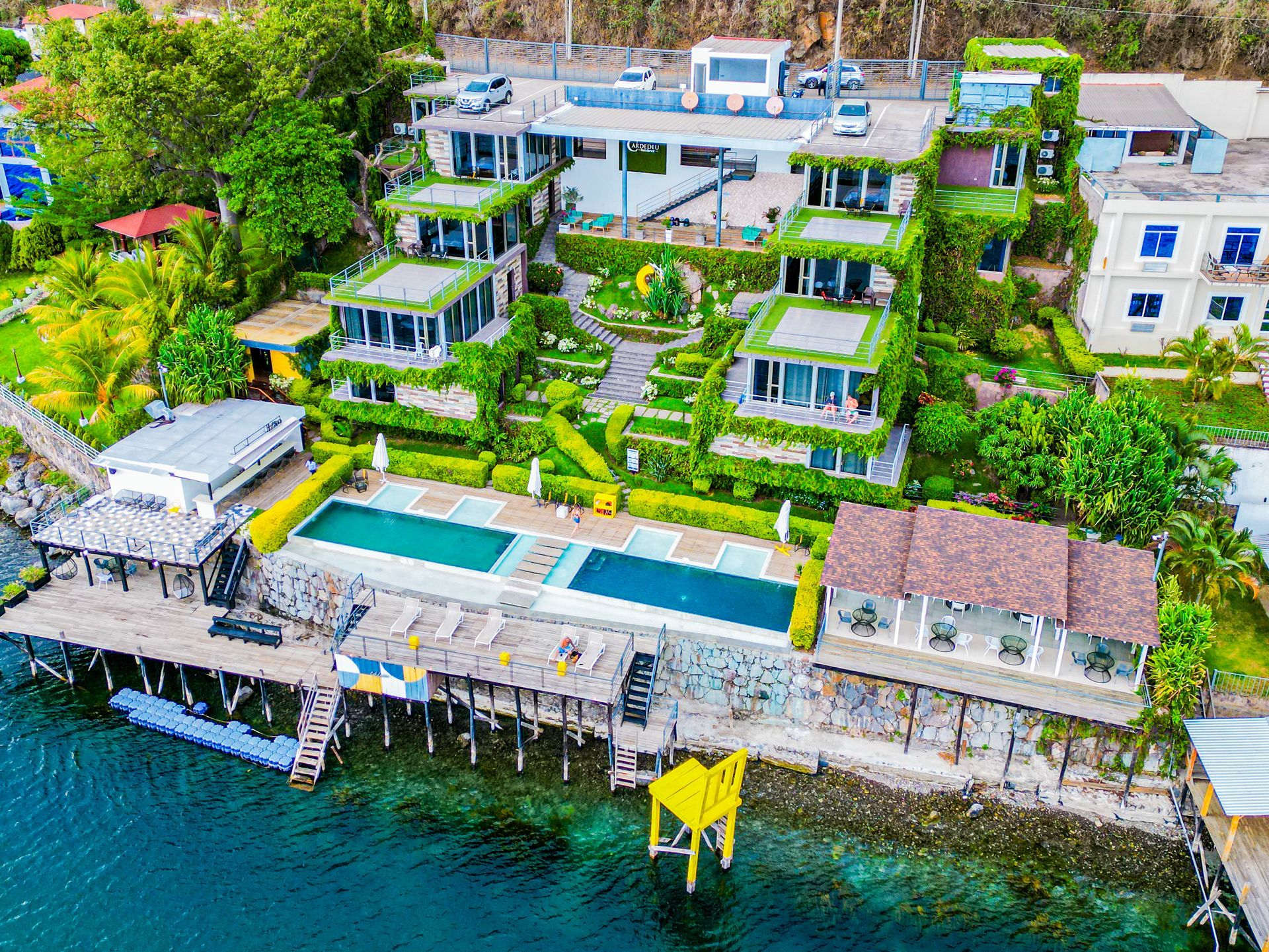 Vista aérea de residence cardedeu coatepeque con villas aterrazadas verdes, piscina y muelle sobre aguas turquesas.