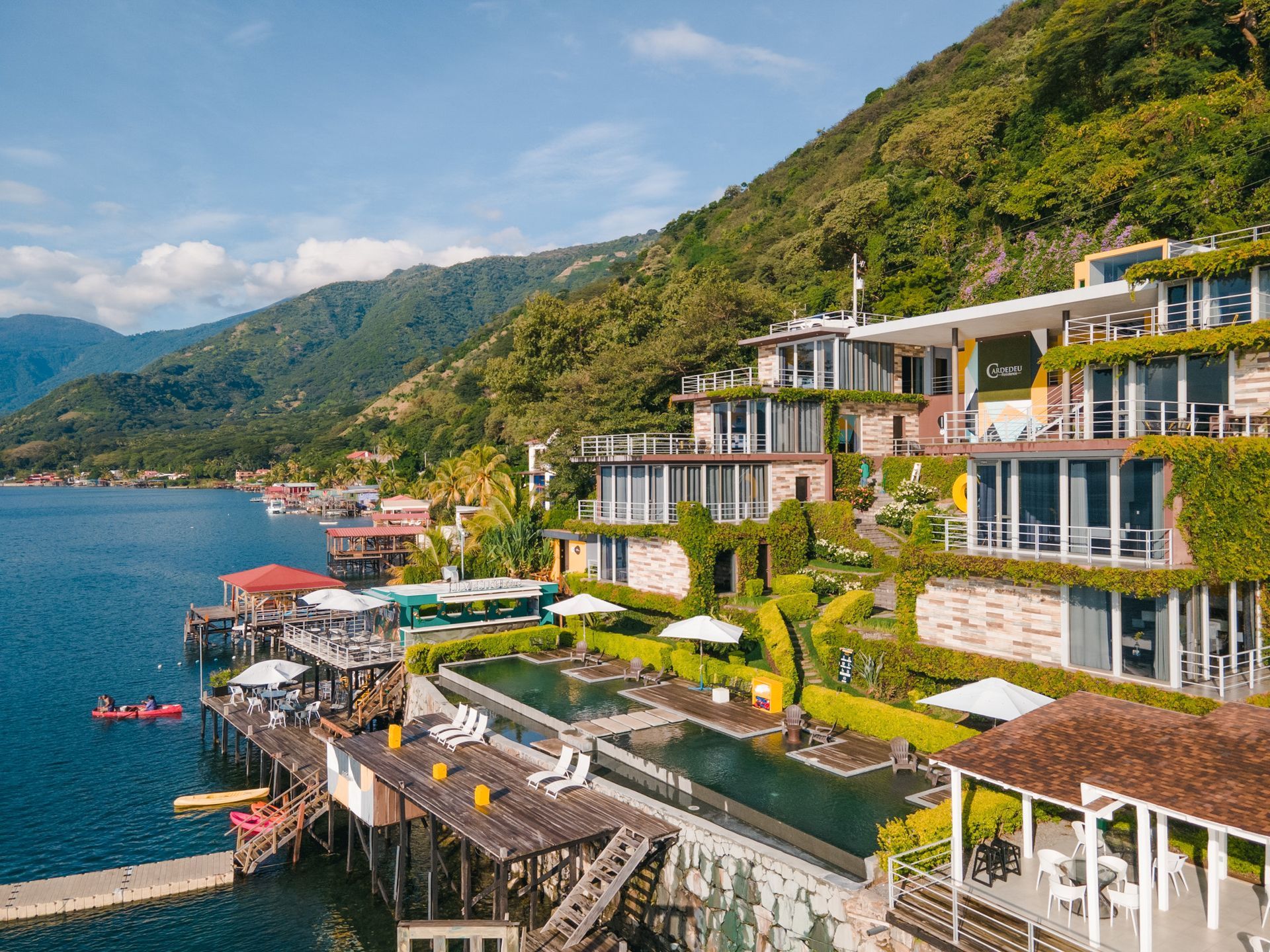 residence cardedeu coatepeque  en la ladera de una colina con edificios blancos con vistas a un lago azul y montañas bajo un cielo soleado.