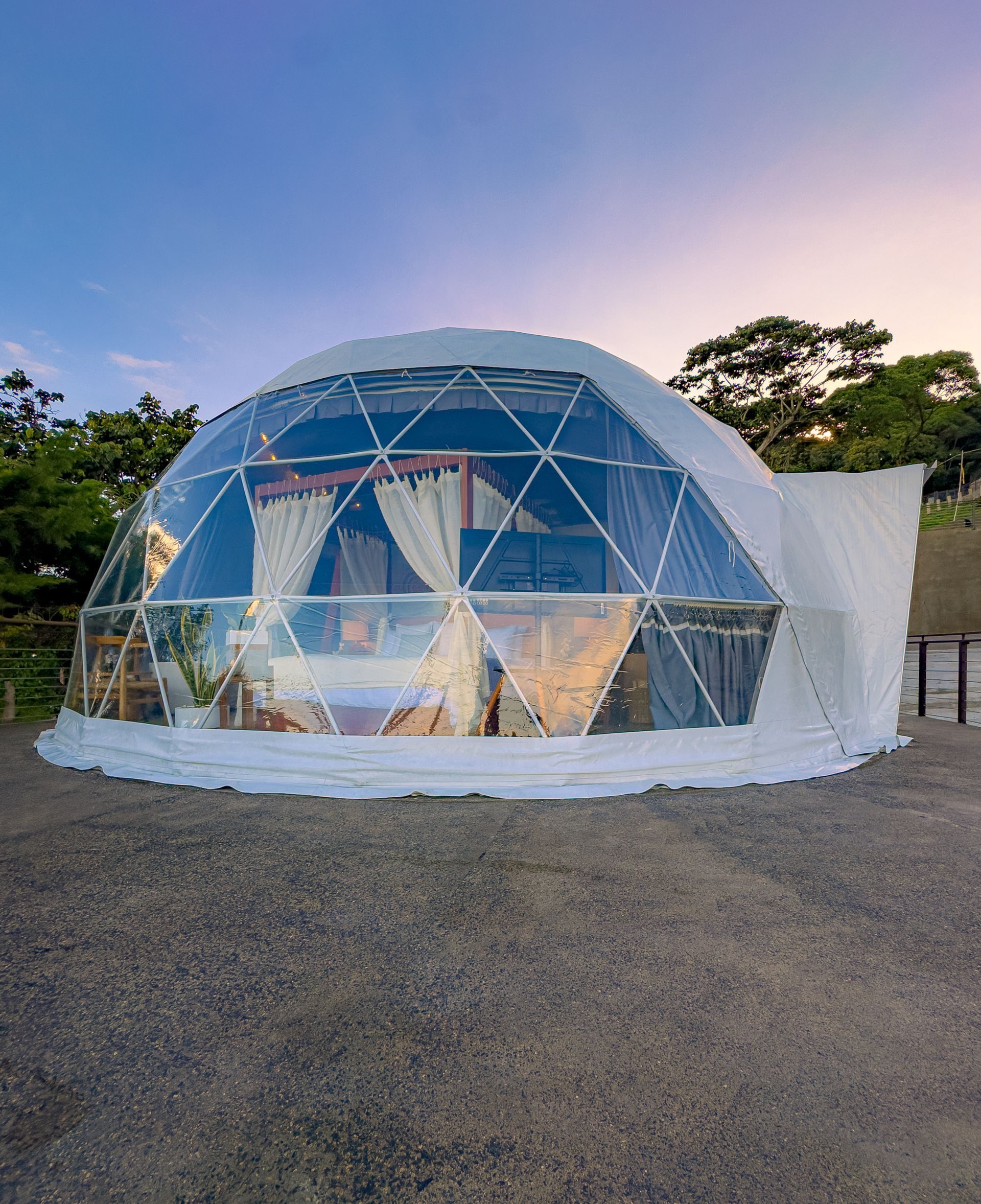 Cabaña con cúpula de cristal geodésica sobre grava al atardecer, con cielo azul y árboles al fondo el boquerón Cardedeu