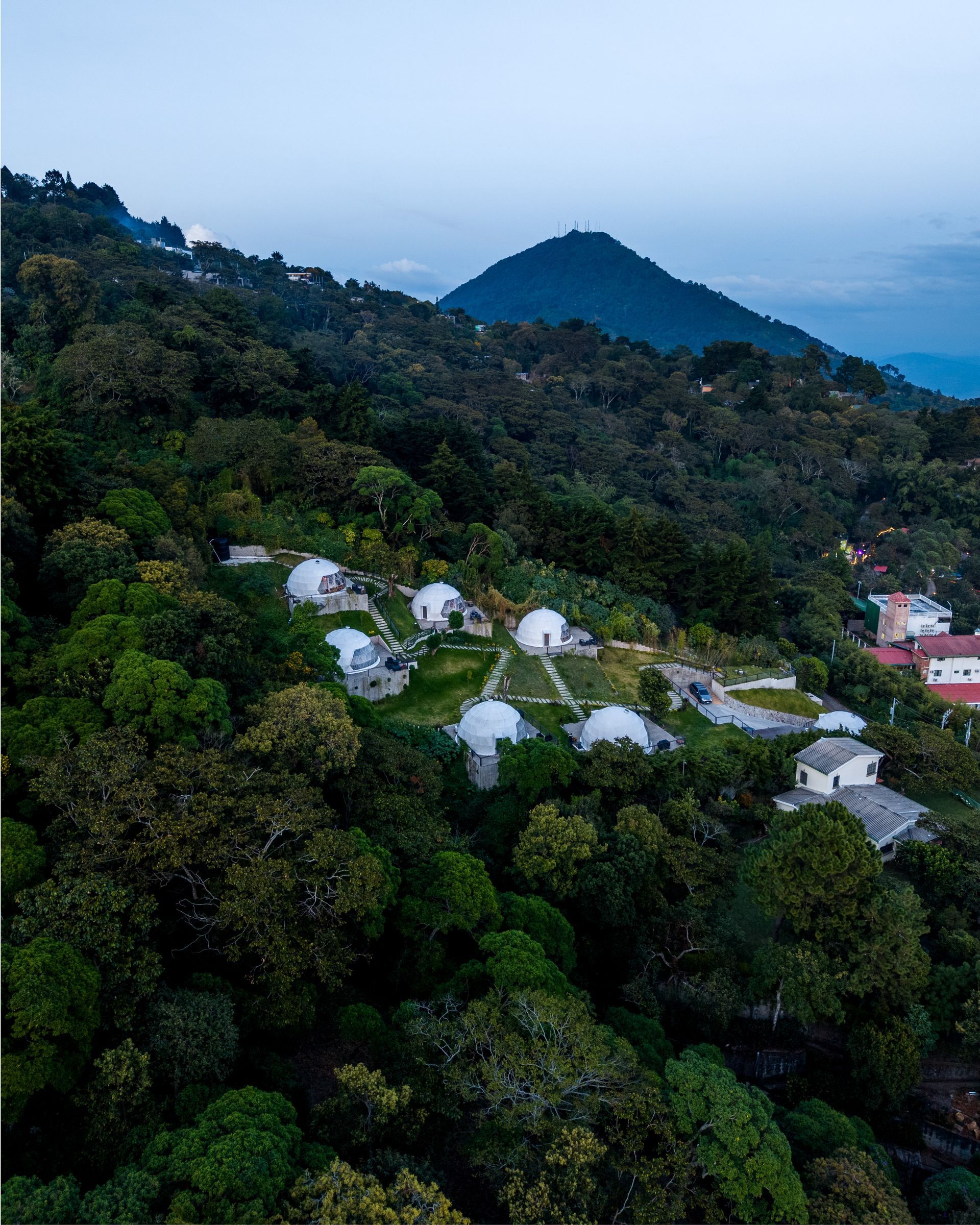 el boquerón Cardedeu en la ladera de una colina, con cabañas blancas enclavadas en un denso bosque verde, con vistas a un mar azul lejano al atardecer.