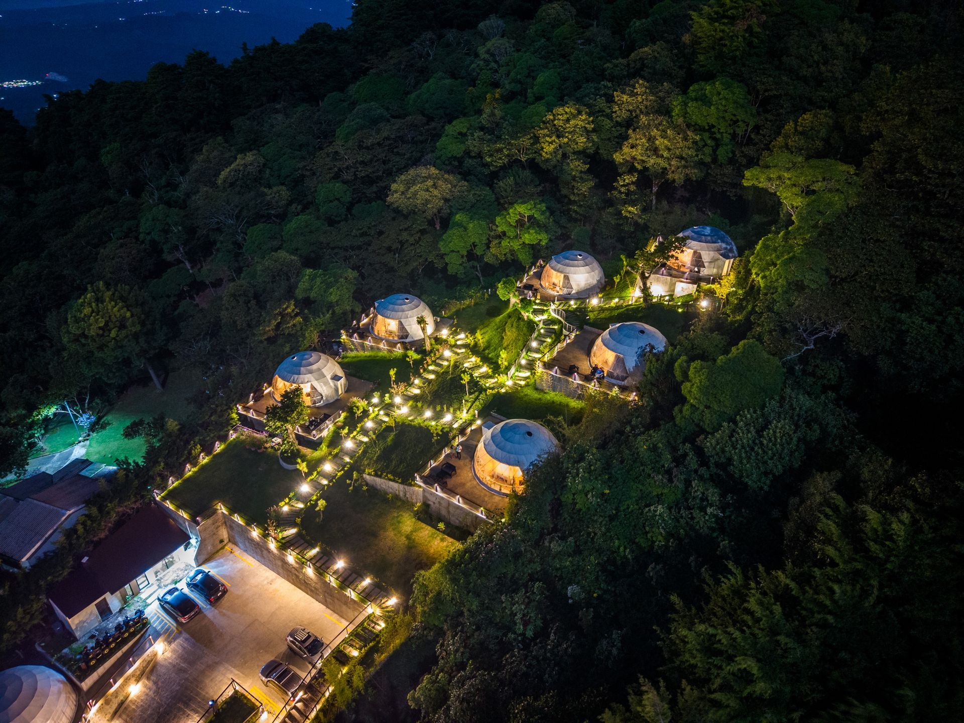 Complejo turístico iluminado en la cima de una colina por la noche, con cabañas redondas, senderos serpenteantes y un área de estacionamiento iluminada en la parte inferior en el boquerón Cardedeu