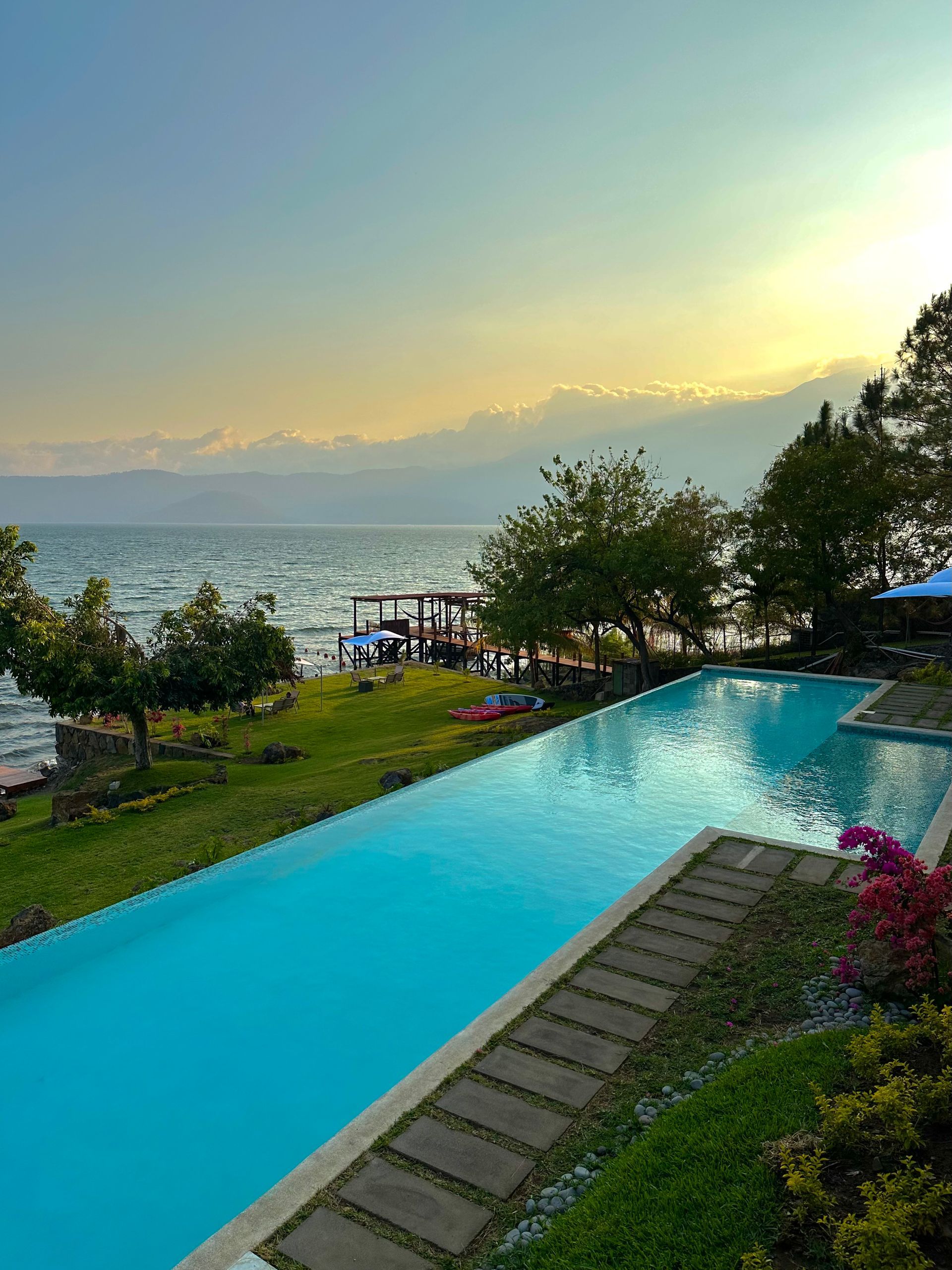 Piscina infinita con vistas al océano al atardecer, con exuberante vegetación y zona de descanso en las inmediaciones.