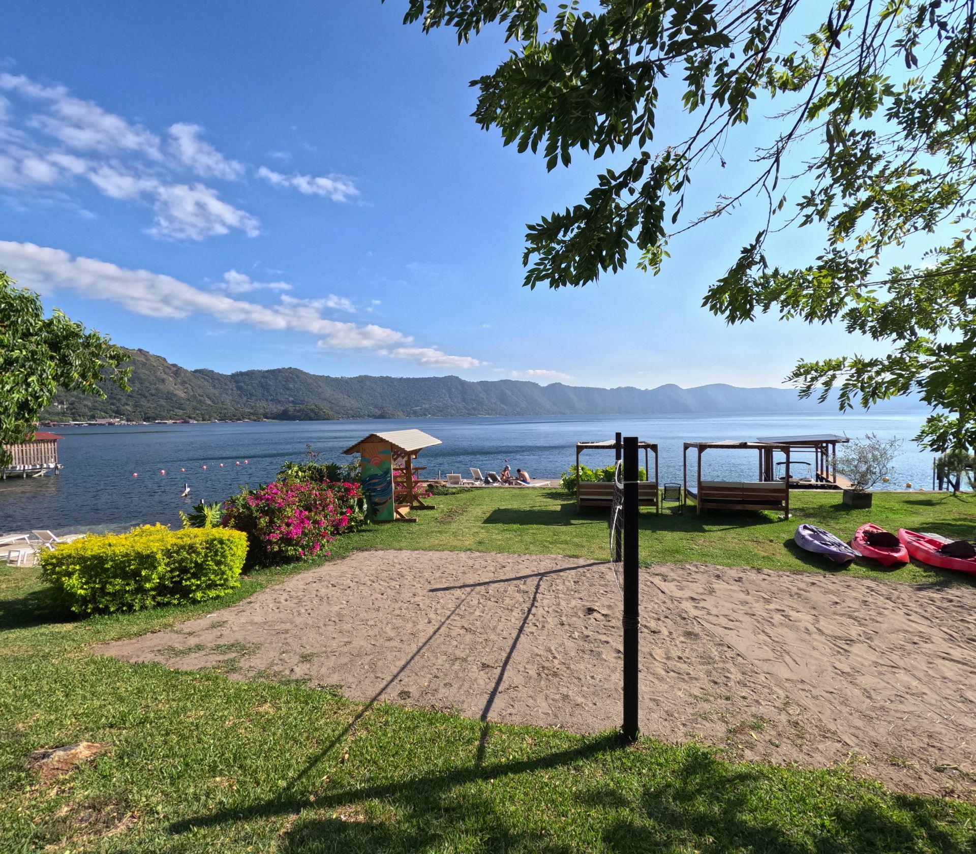 Playa soleada a orillas del lago coatepeque con ribera cubierta de hierba, mesas de picnic, sombrillas y kayaks bajo un cielo azul.