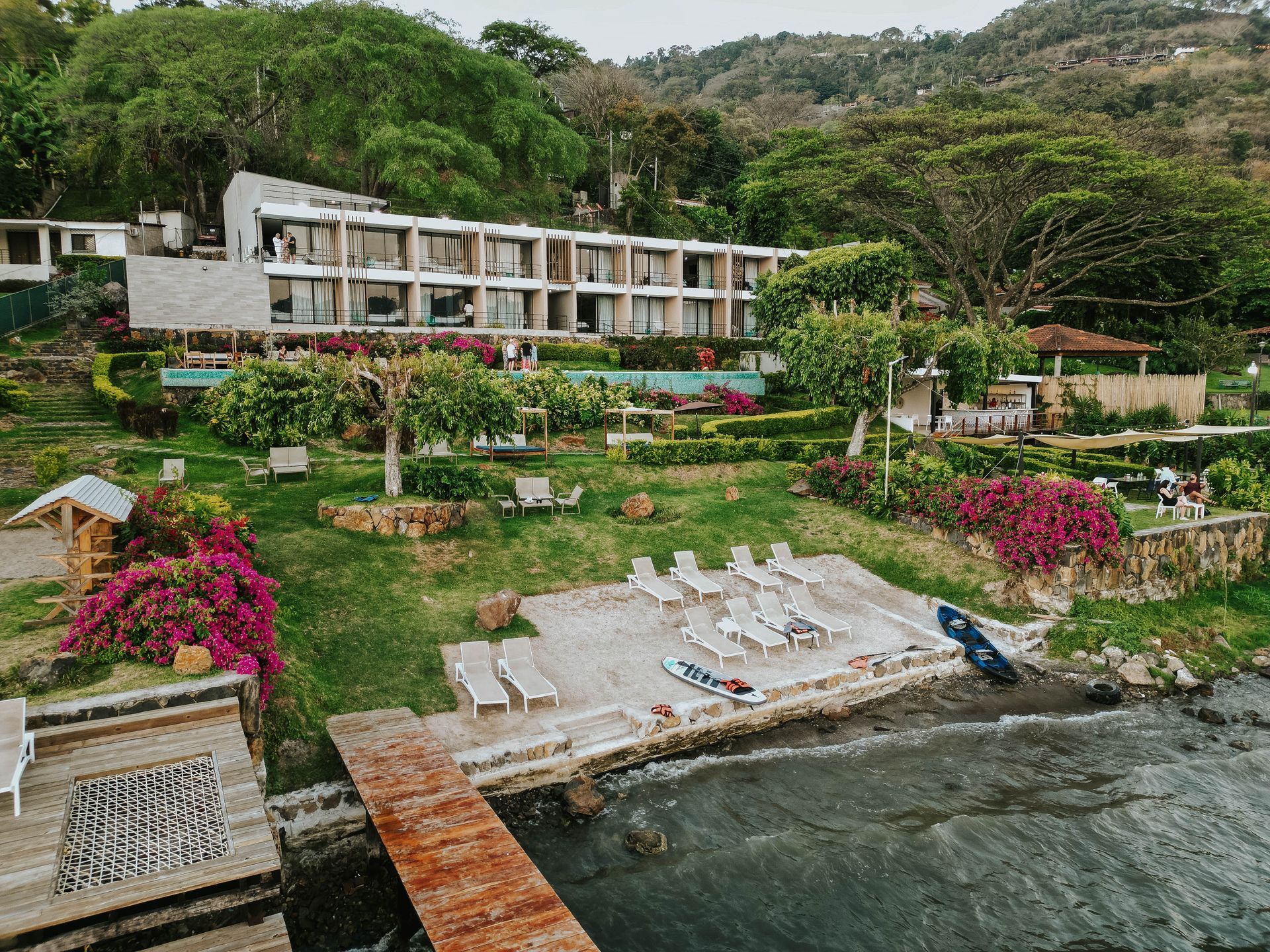 hotel cardedeu coatepeque con vista al lago edificios blancos, jardines exuberantes y tumbonas junto a una costa rocosa.