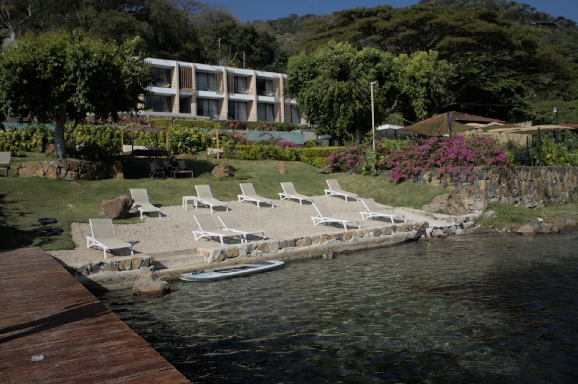 hotel cardedeu coatepeque con tumbonas junto al agua, jardines tropicales y un hotel moderno al fondo.