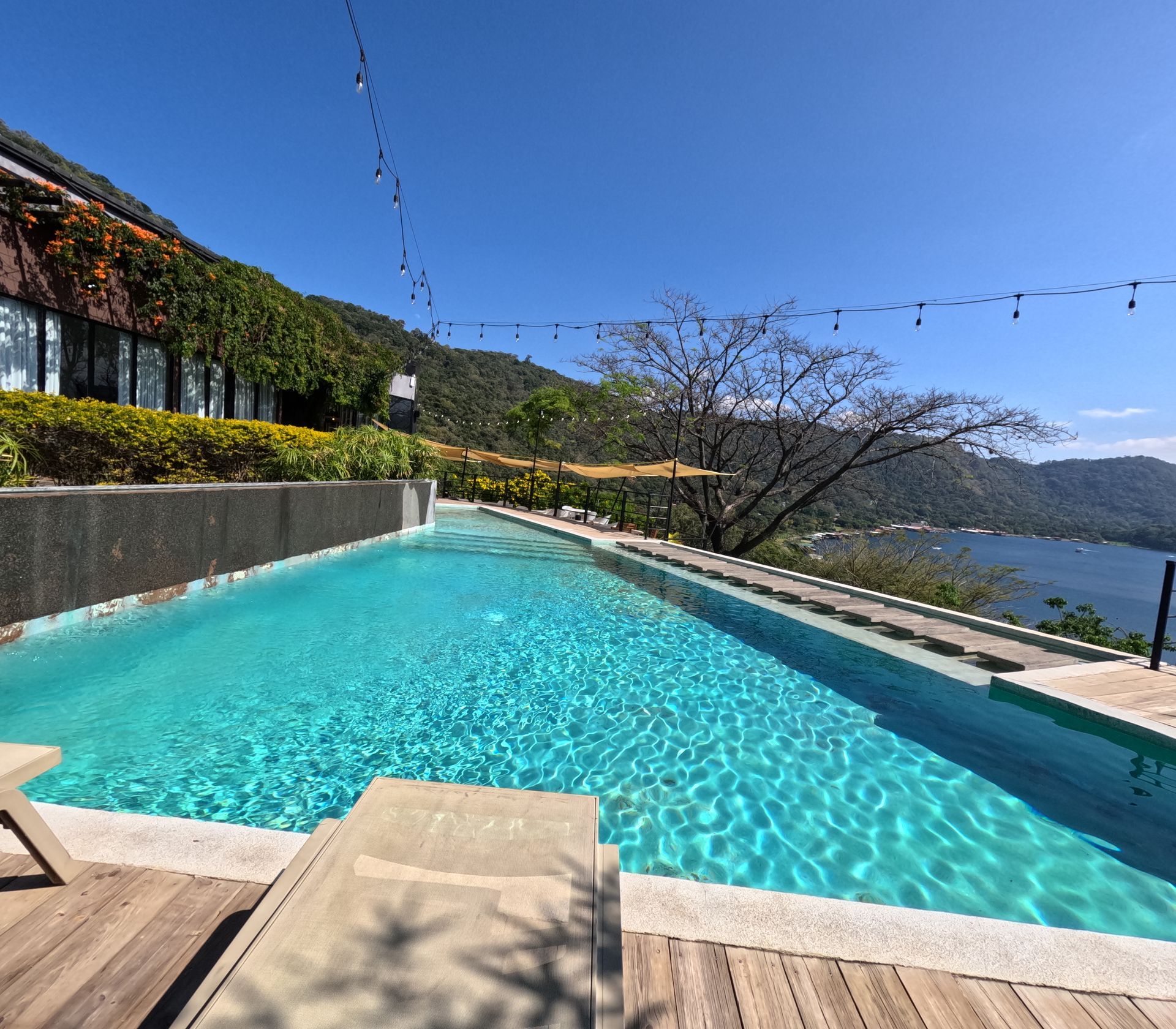 Piscina infinita con agua turquesa con vistas a un lago y colinas bajo un cielo azul despejado.