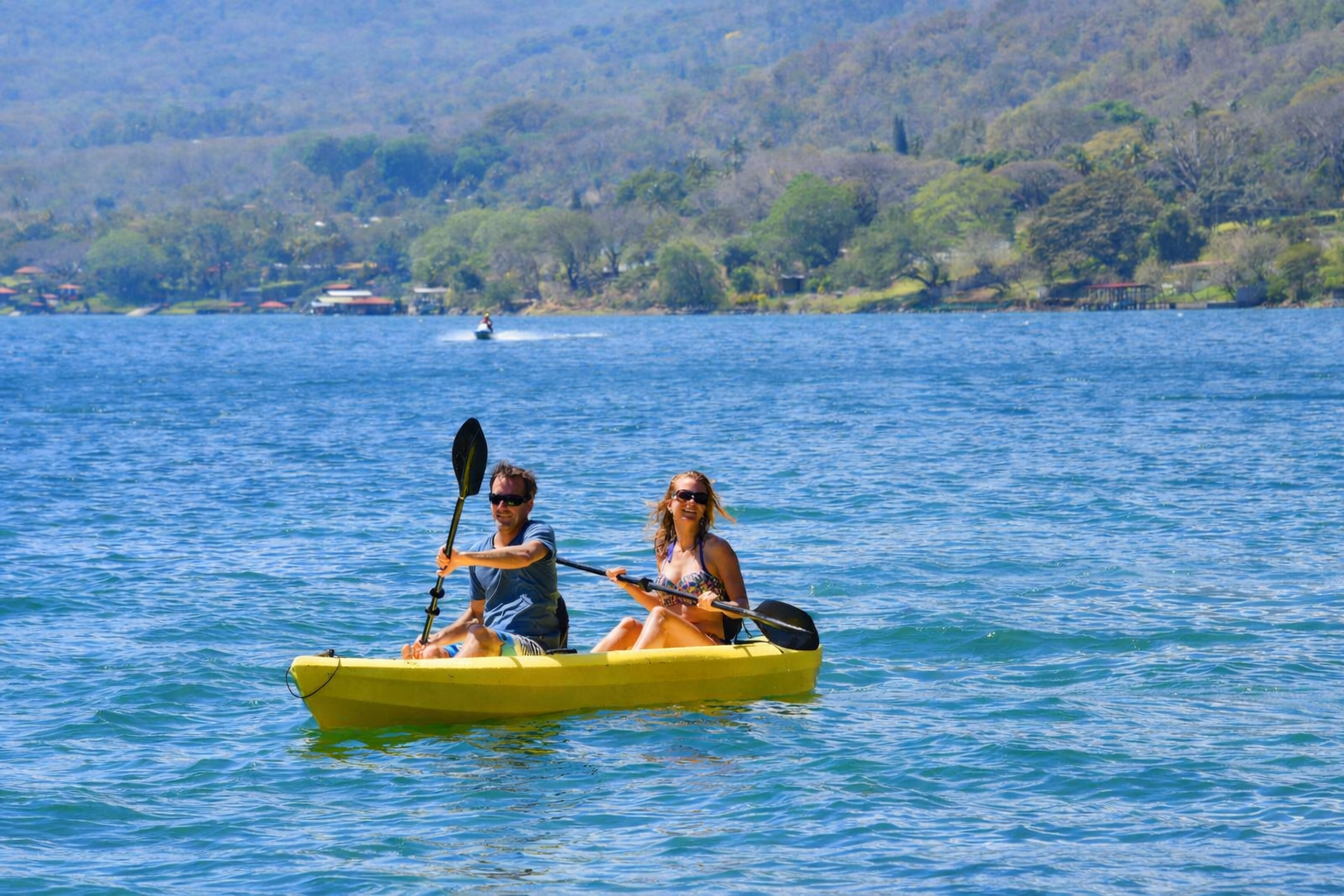 Dos personas remando en un kayak amarillo sobre agua azul con colinas verdes al fondo.