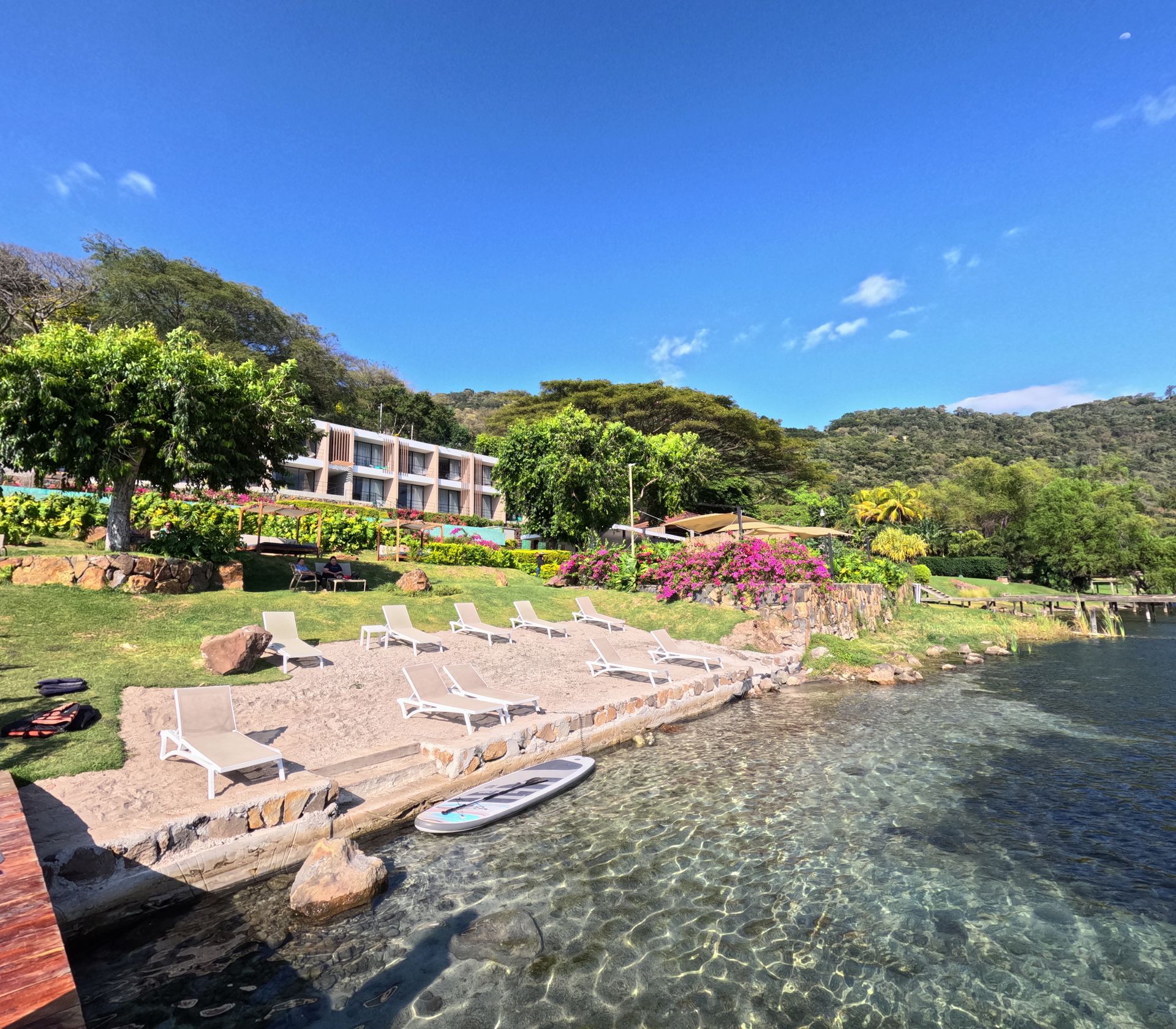 Complejo turístico tropical con tumbonas, aguas cristalinas, flores rosas y vegetación en la ladera bajo un cielo azul en hotel cardedeu coatepeque