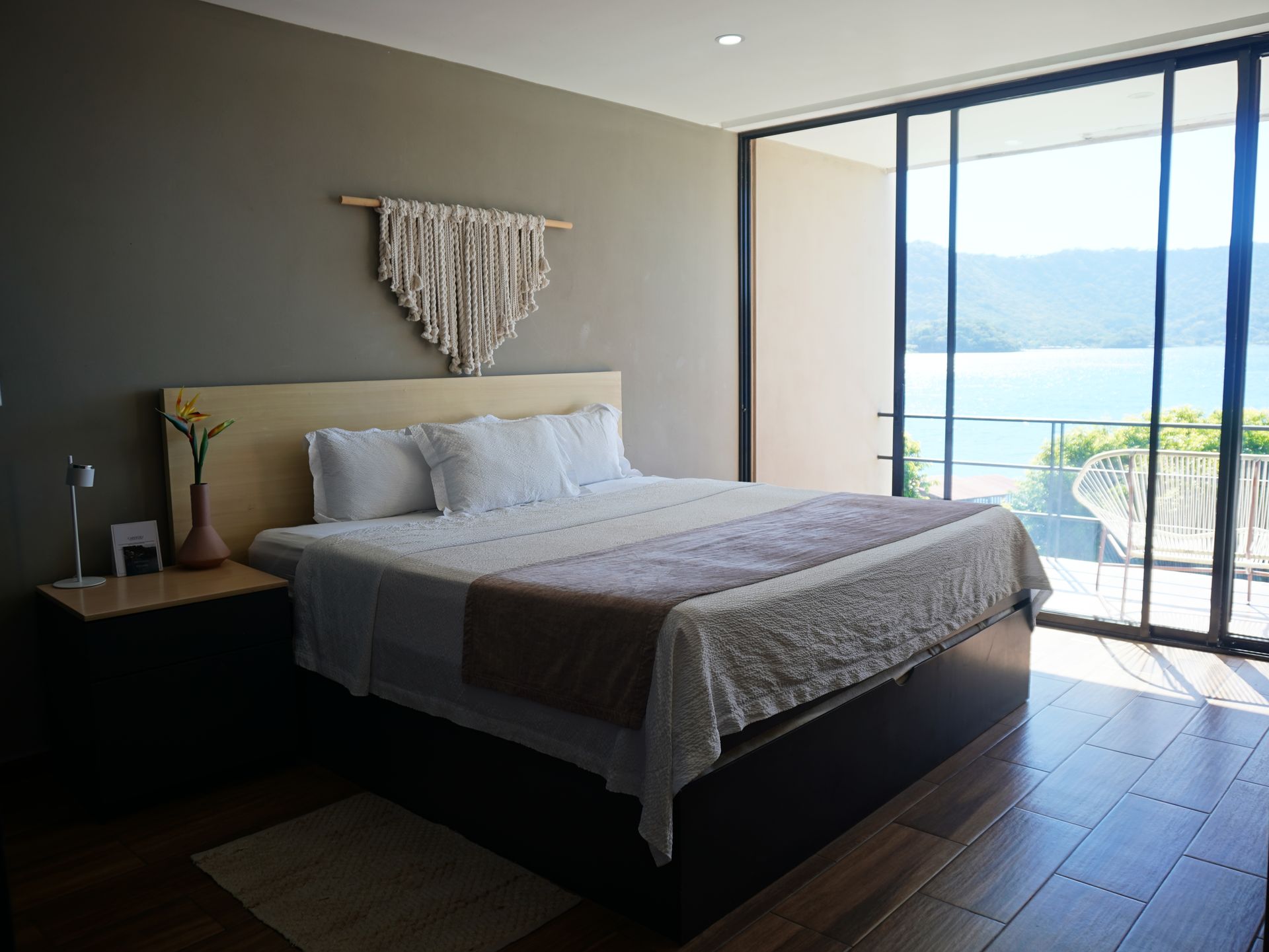 Habitación moderna con cama, mesita de noche y balcón con vistas a un lago y montañas en el hotel cardedeu coatepeque