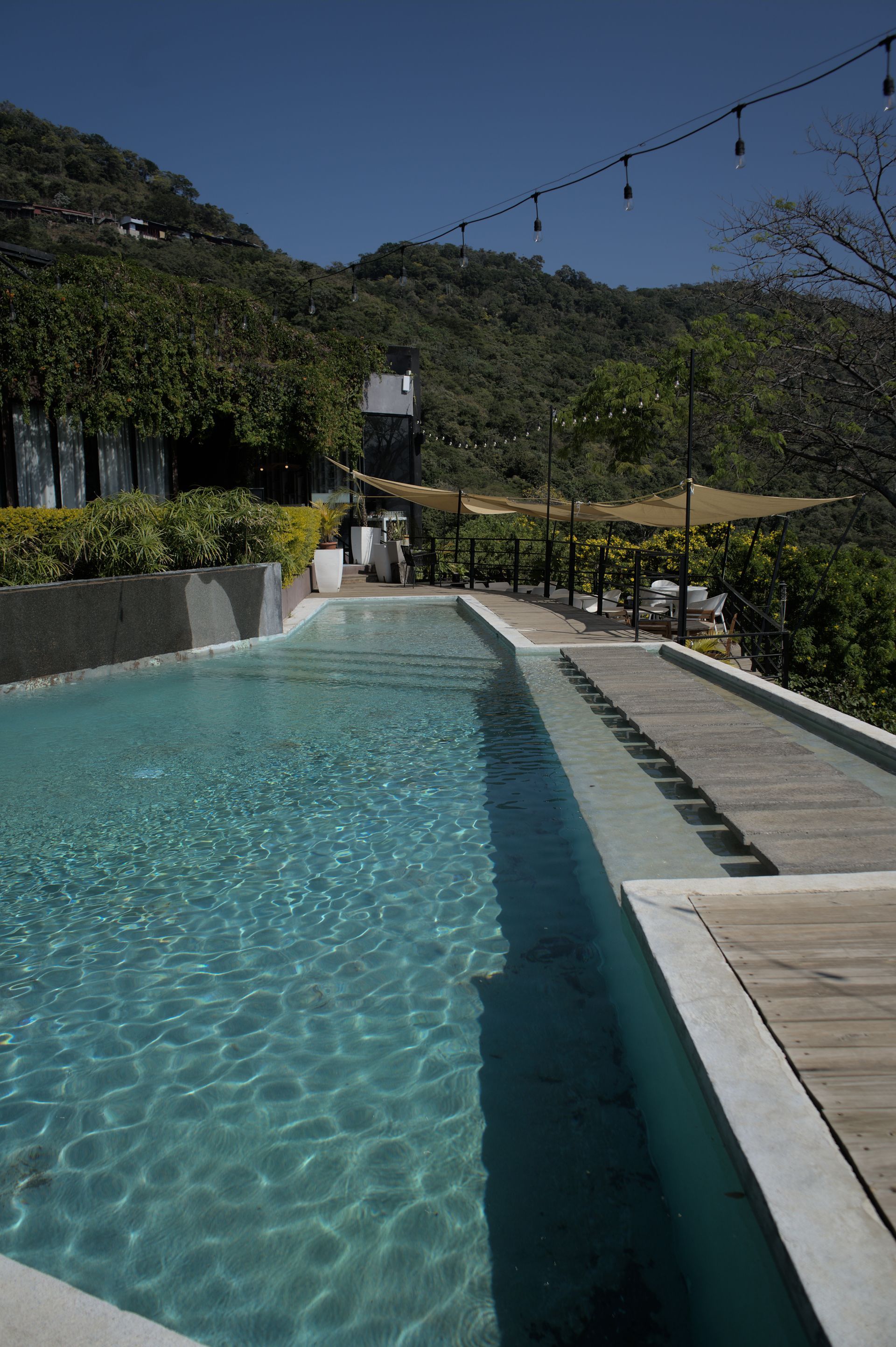 Piscina infinita junto a una pasarela de madera, con árboles, colinas y luces de guirnalda al fondo.
