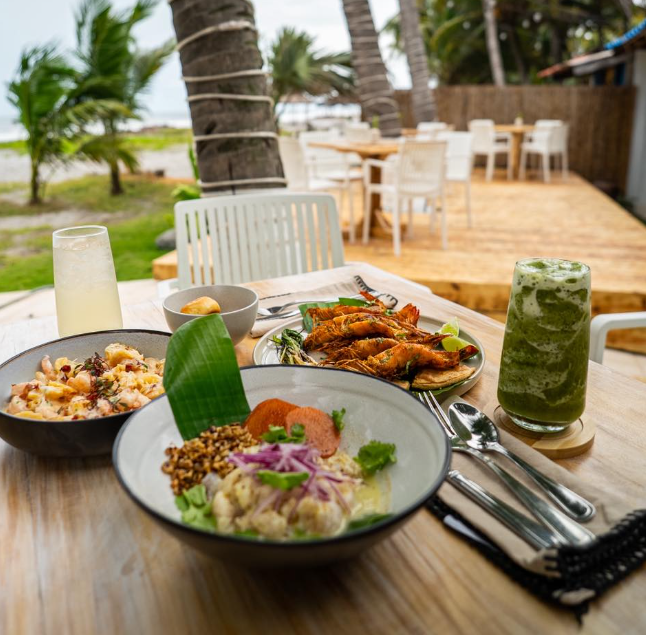 Mesa en restaurante Nacar junto a la playa con platos tropicales y una bebida verde en un patio de madera junto a las palmeras.
