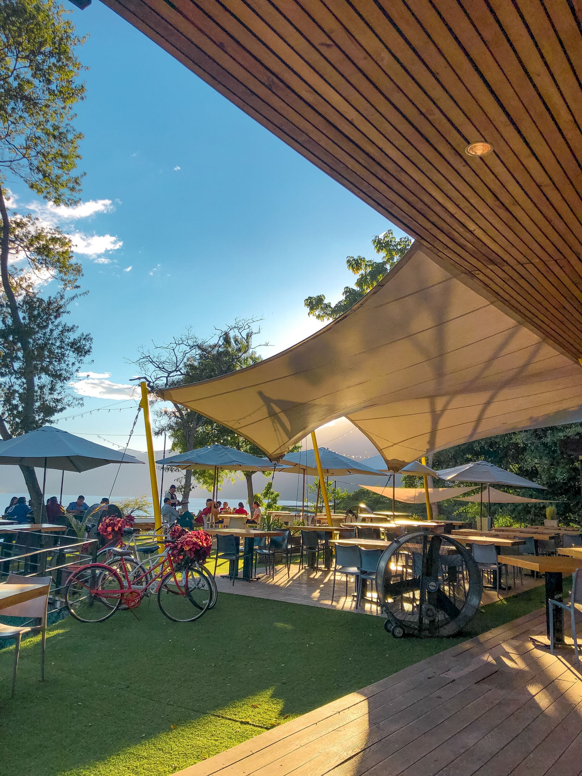 Terraza de cafetería al aire libre con toldos blancos, bicicletas, mesas y césped verde bajo un pérgola de madera.