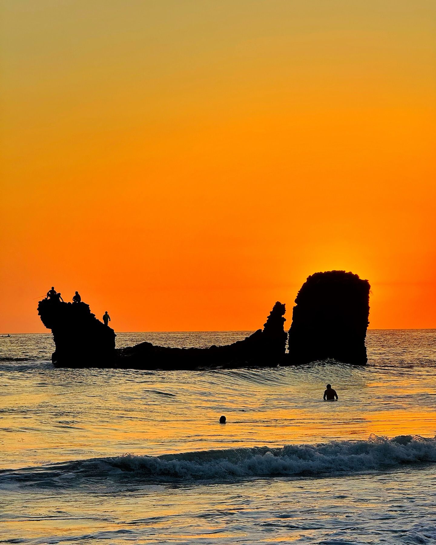 Silueta de una formación rocosa en el mar al atardecer, con cielo anaranjado y olas suaves.