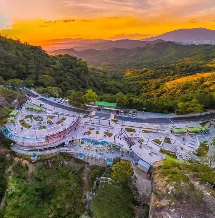 Vista del atardecer desde un mirador de montaña curvado con terrazas, vegetación y colinas distantes bañadas por una cálida luz dorada.