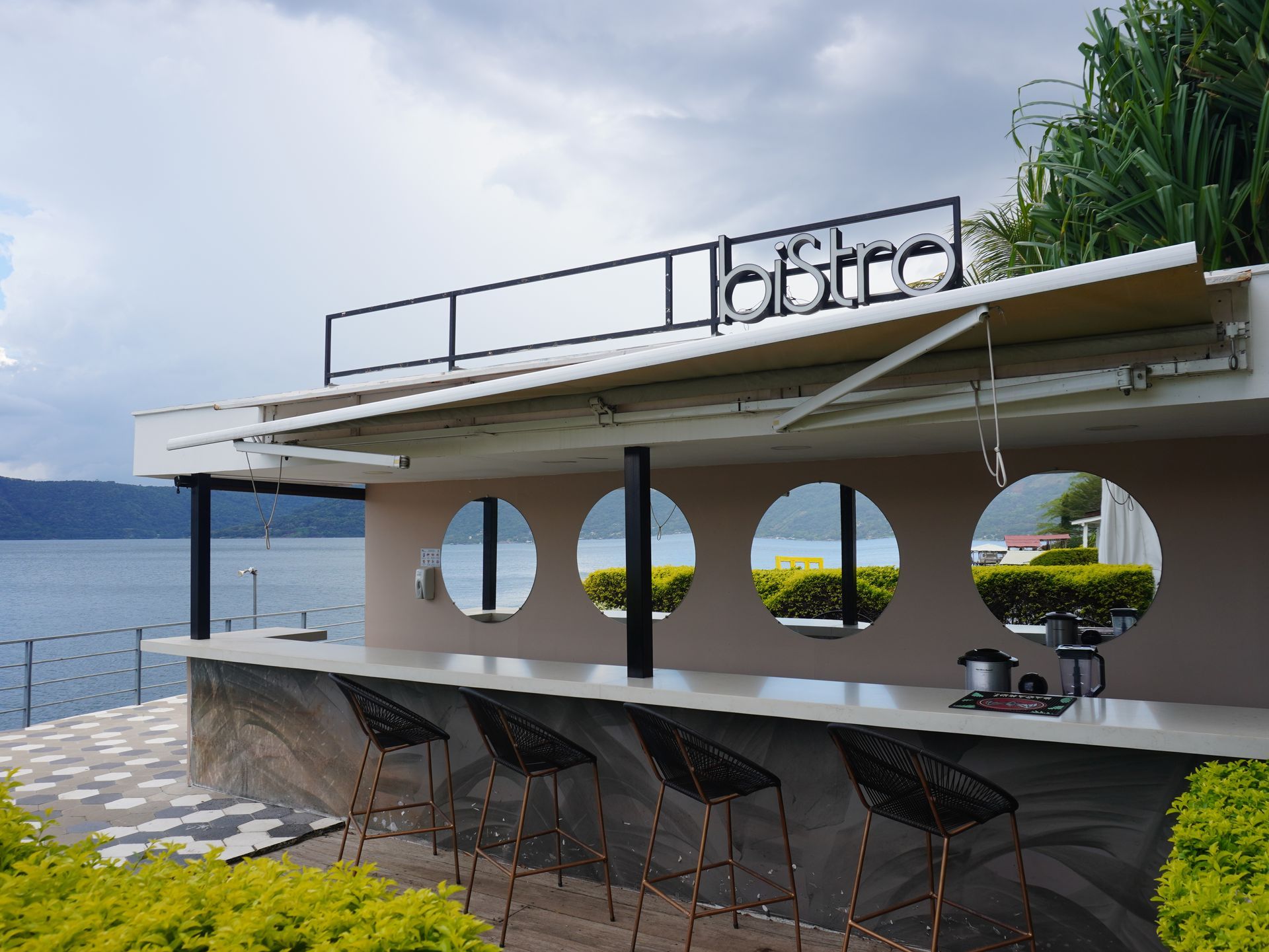Bar moderno frente al mar con ventanas circulares, taburetes y vistas al océano bajo un cielo nublado.