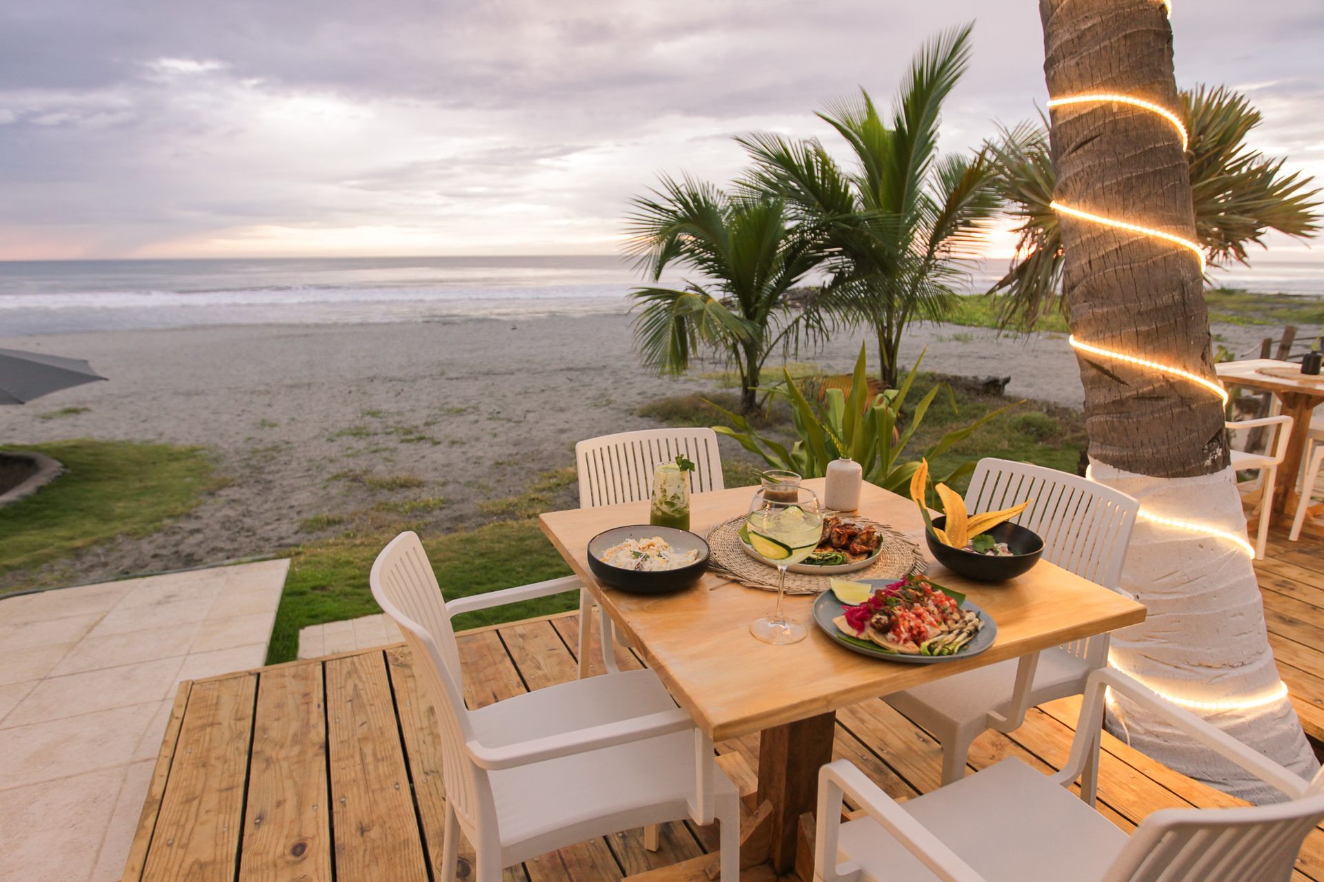 Mesa de comedor en un patio junto al mar con decoración tropical, palmera iluminada y vista al océano al atardecer.