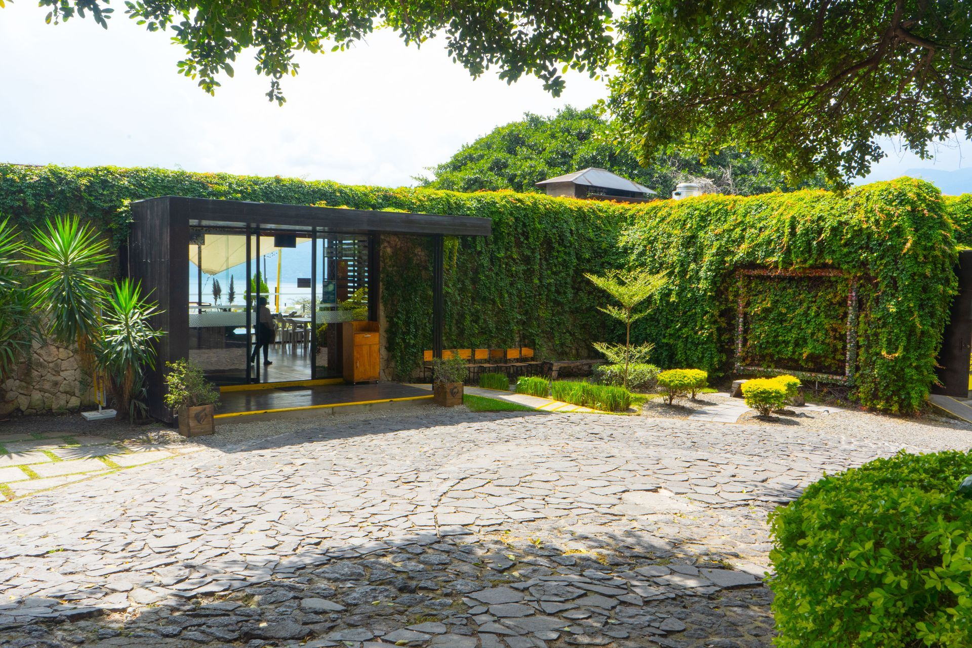 Patio de piedra frente a un moderno edificio cubierto de hiedra con puertas de cristal y zonas verdes ajardinadas en pampa restaurante hotel cardedeu coatepeque