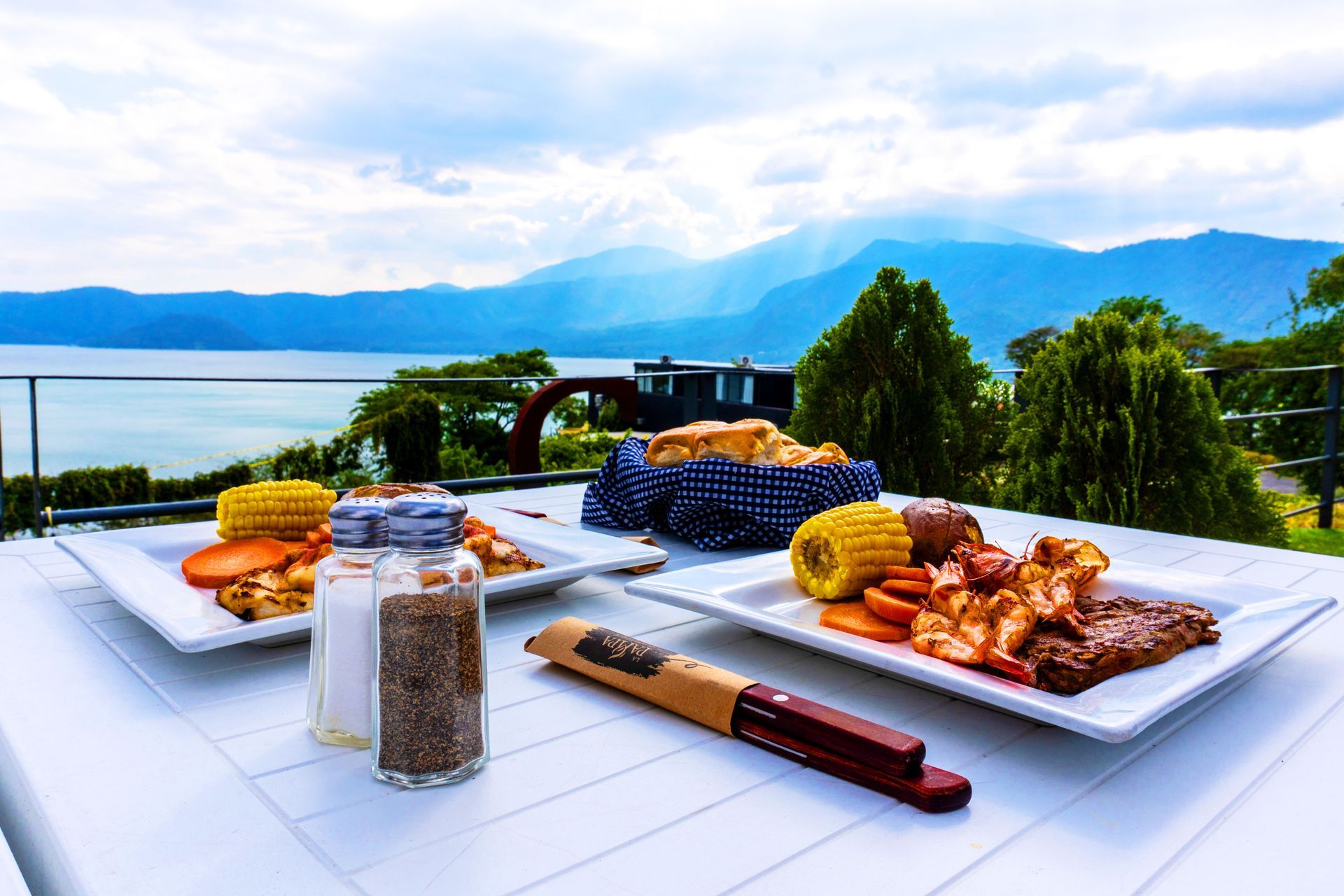 Mesa al aire libre con comida a la parrilla, pan y bebidas con vistas a un lago y a las montañas  en restaurante pampa hotel coatepeque